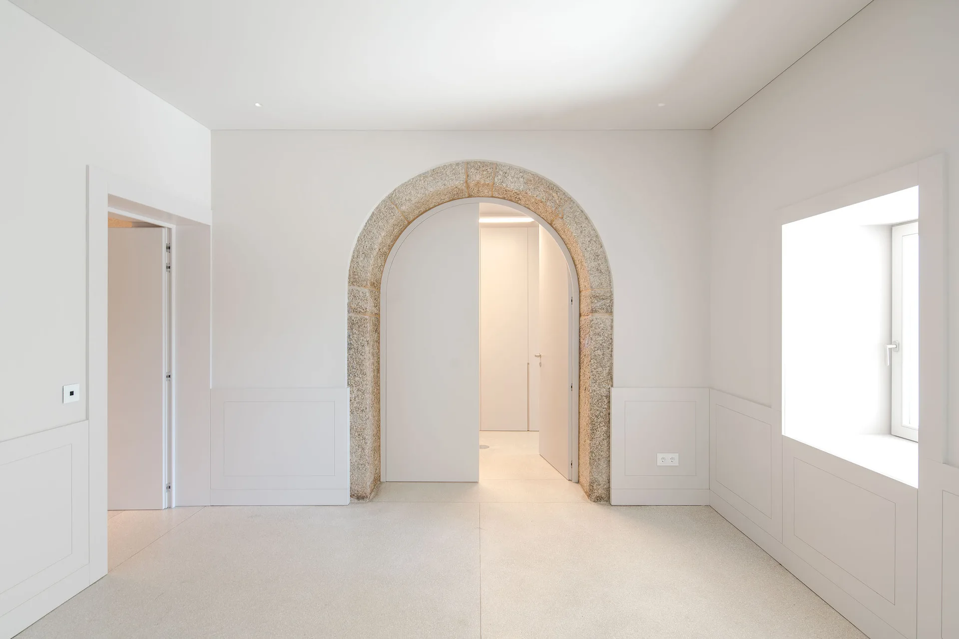 A minimalist white hallway featuring a central stone arched doorway with white doors, lower wall wainscoting, and a recessed window.