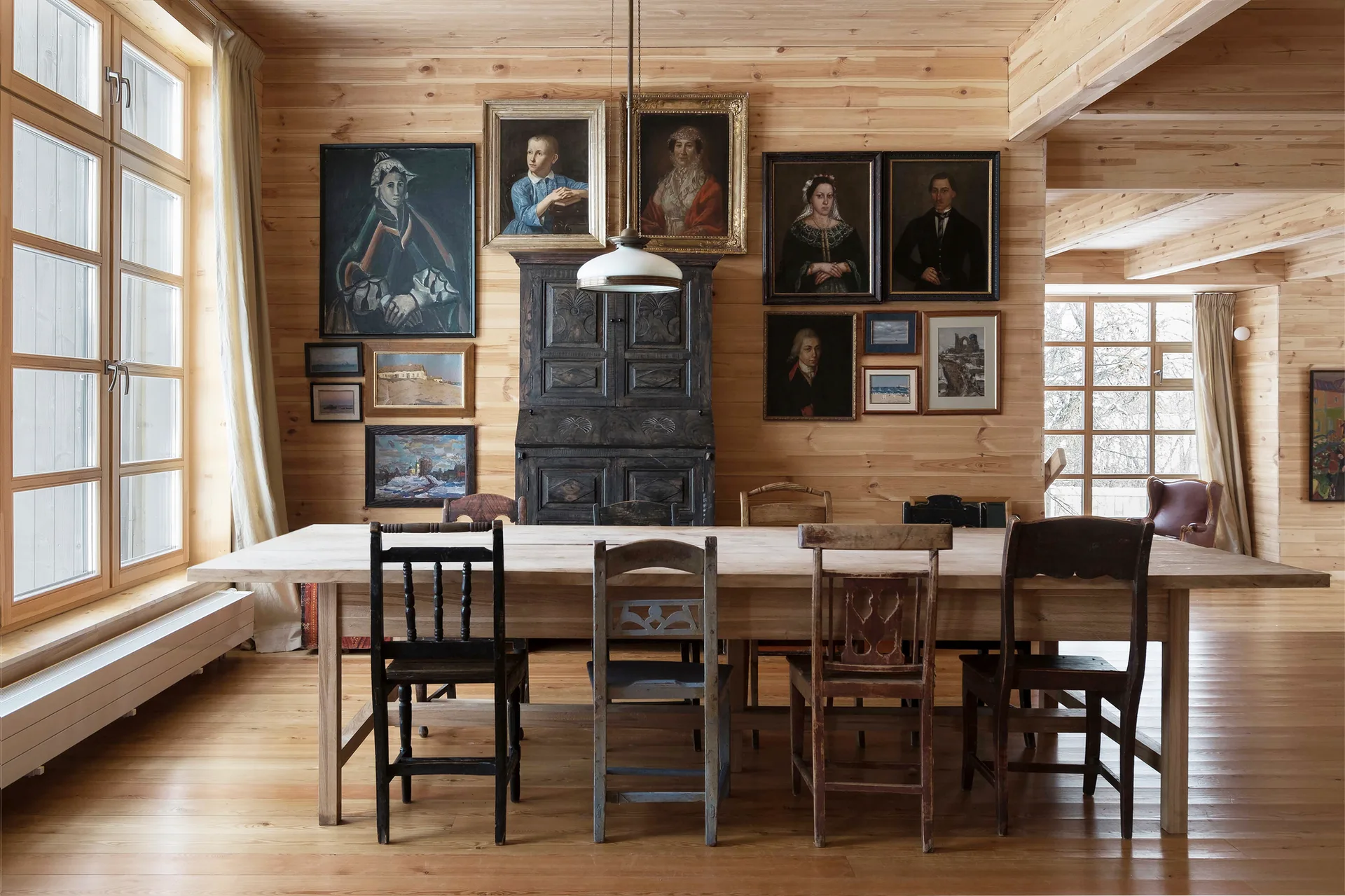 A rustic dining room features a long wood table surrounded by mismatched vintage chairs against a wood-paneled wall decorated with framed portraits.