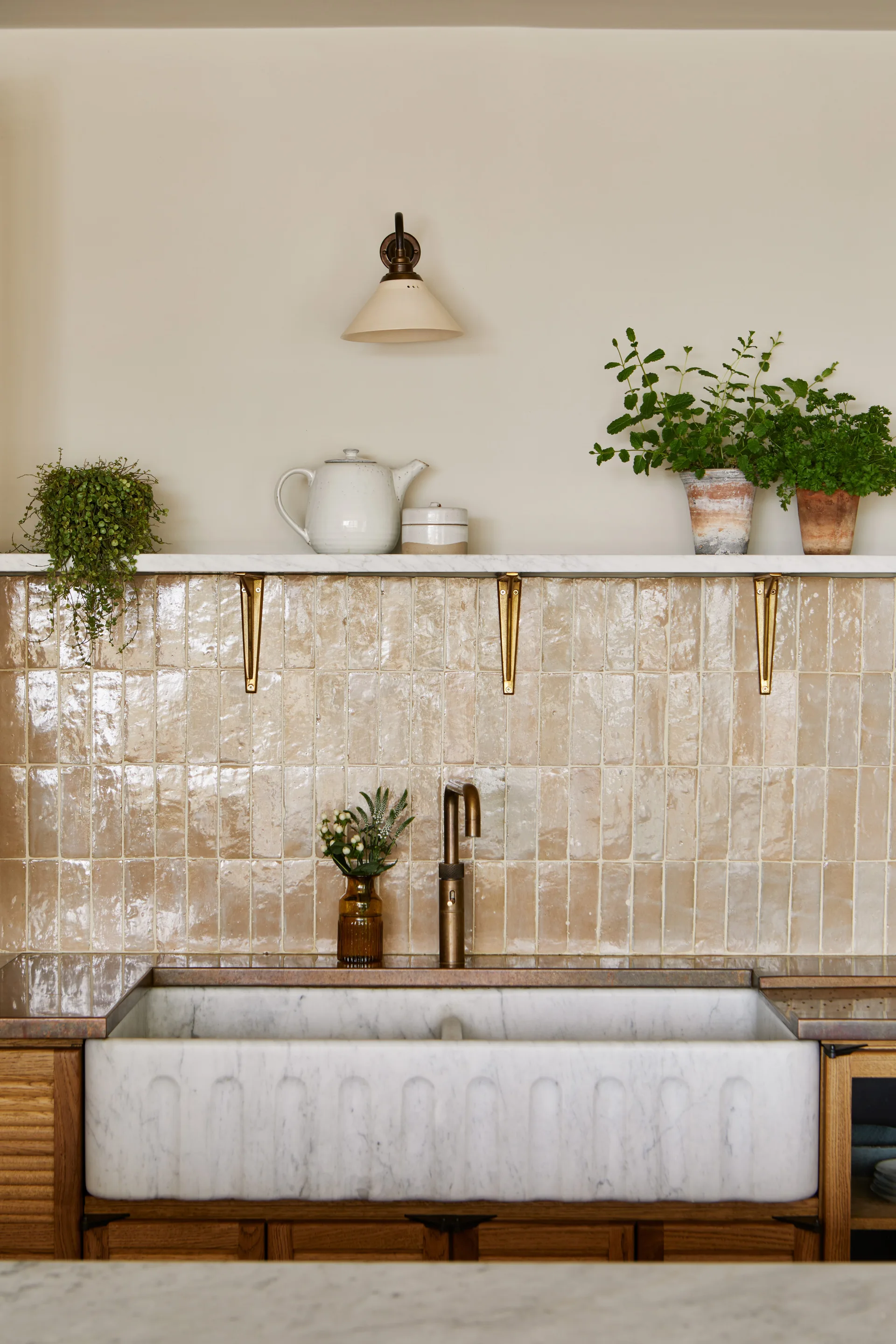 A white marble farmhouse sink features a fluted front beneath a glossy beige tiled backsplash, brass faucet, and a stone shelf holding potted plants.