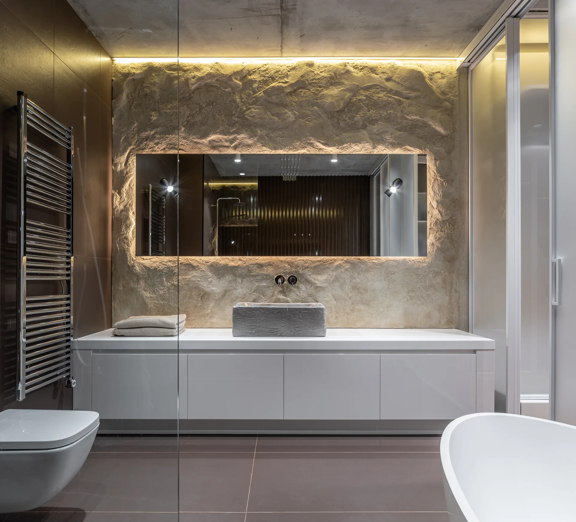 A modern bathroom with a white floating vanity, a stone vessel sink, and a large backlit mirror mounted on a rough textured rock wall.
