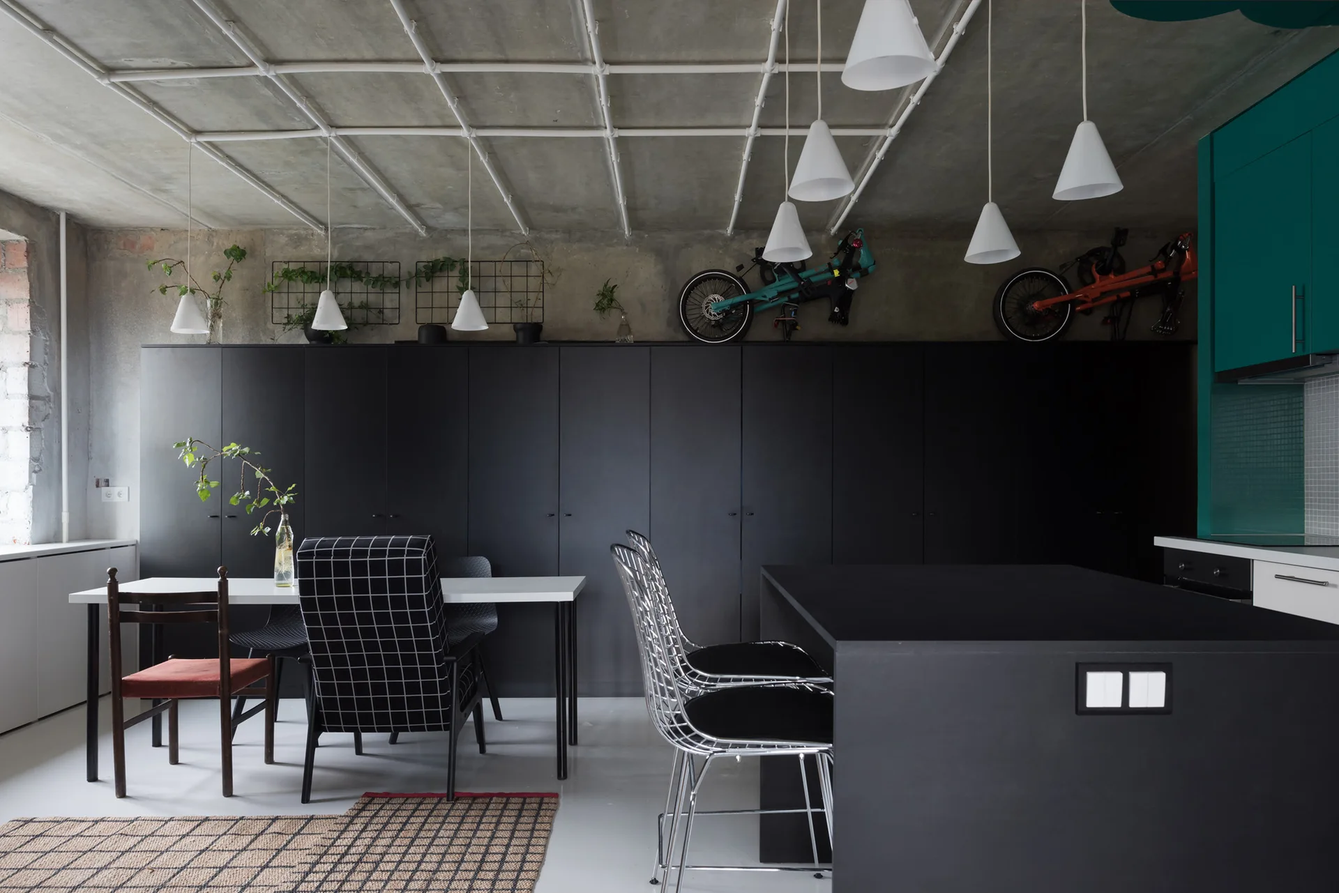 An industrial kitchen and dining area featuring matte black cabinets, an exposed concrete ceiling with white grid pipes, and cone pendant lights.
