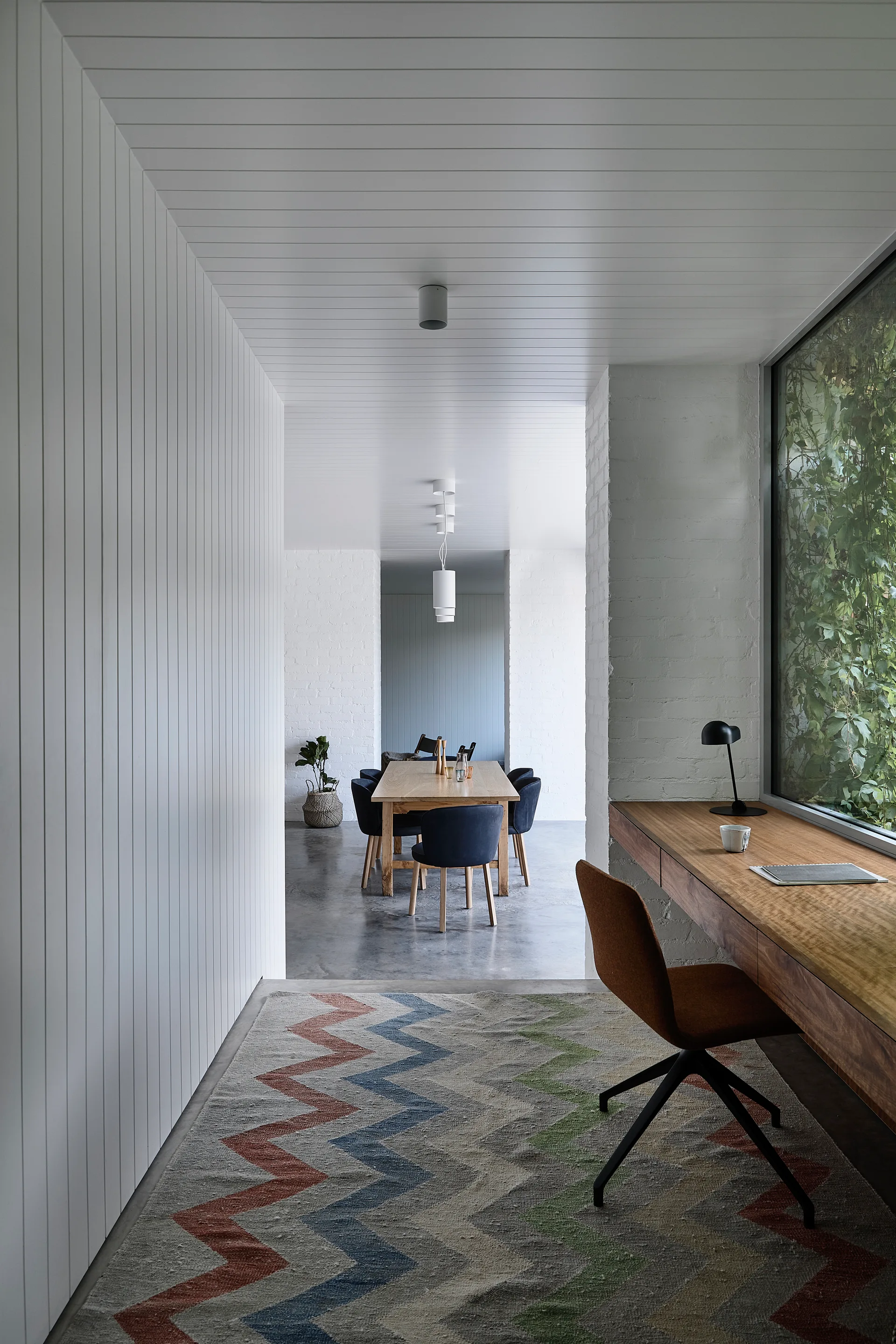 A long wooden desk sits beneath a window in a modern home office with vertical white wall paneling, a chevron rug, and concrete floors.