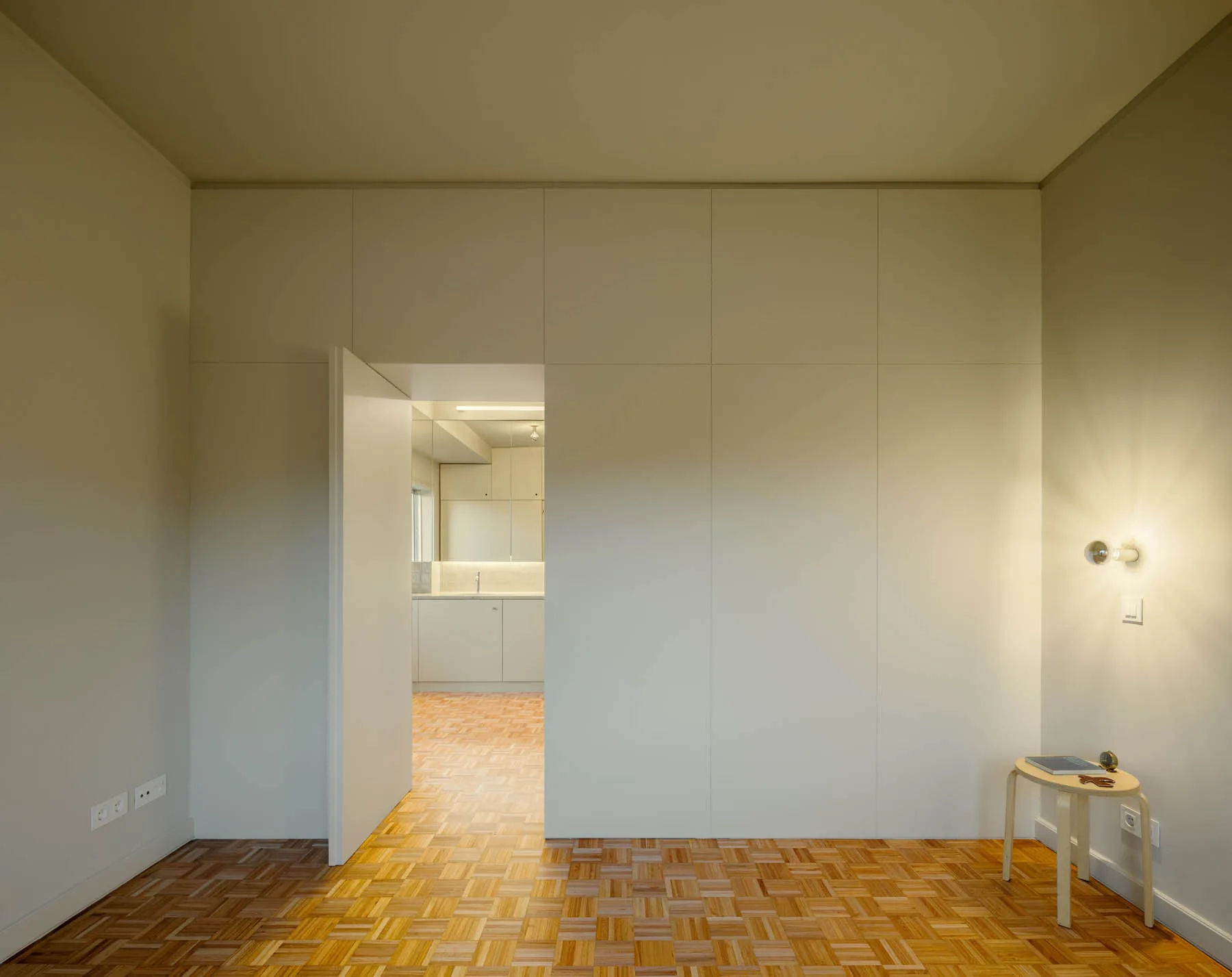 An empty room features an integrated off-white cabinetry wall, an open doorway, a wooden stool, and geometric parquet flooring.