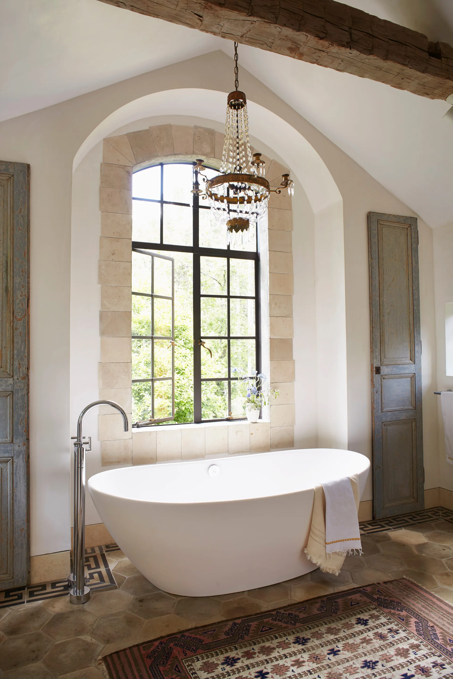 A white freestanding bathtub sits on a hexagonal tile floor beneath an arched window with stone trim and a crystal chandelier.