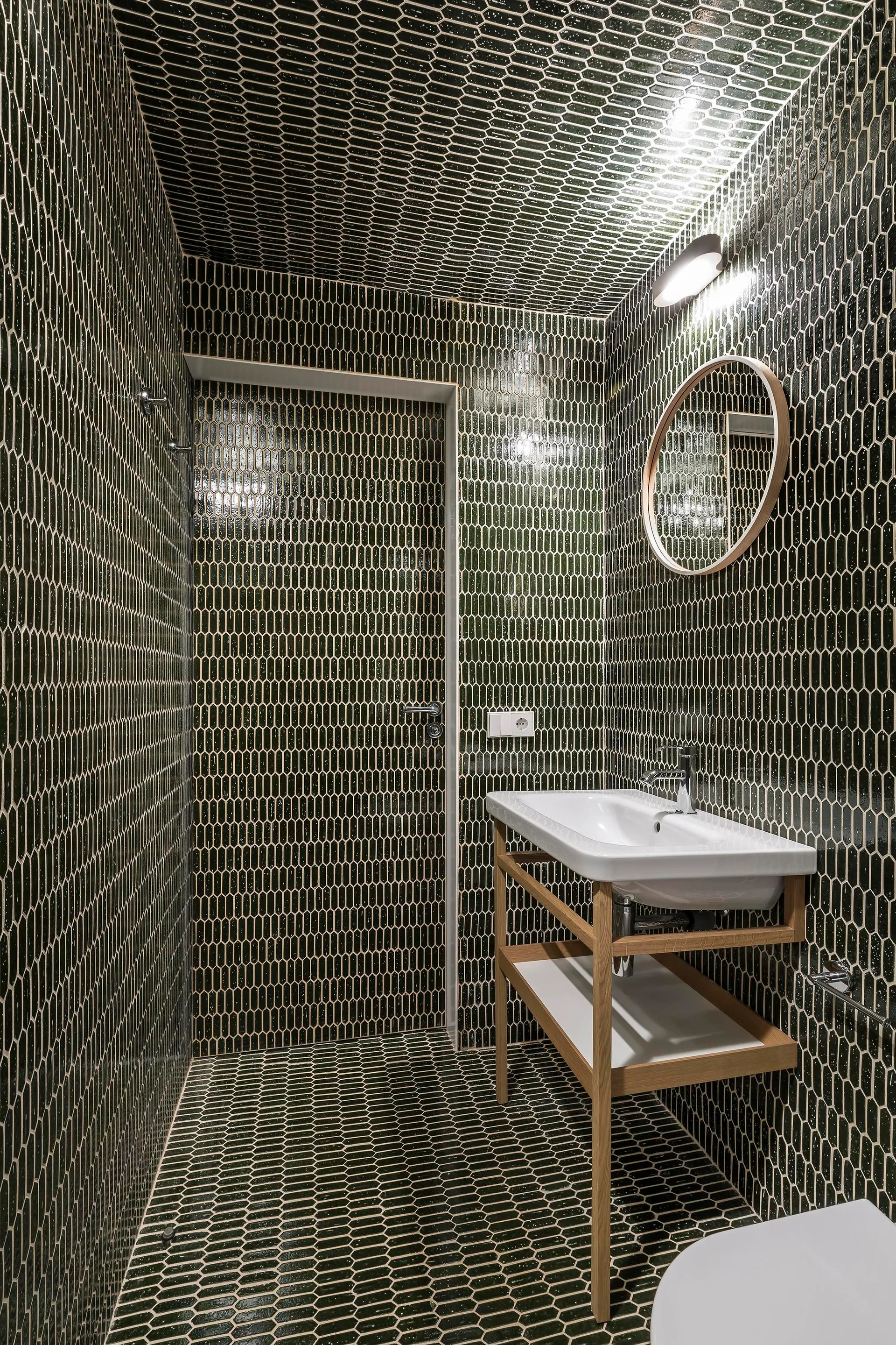 A modern bathroom featuring glossy dark green picket tiles on the walls, floor, and ceiling with a white sink on a light wood frame vanity.