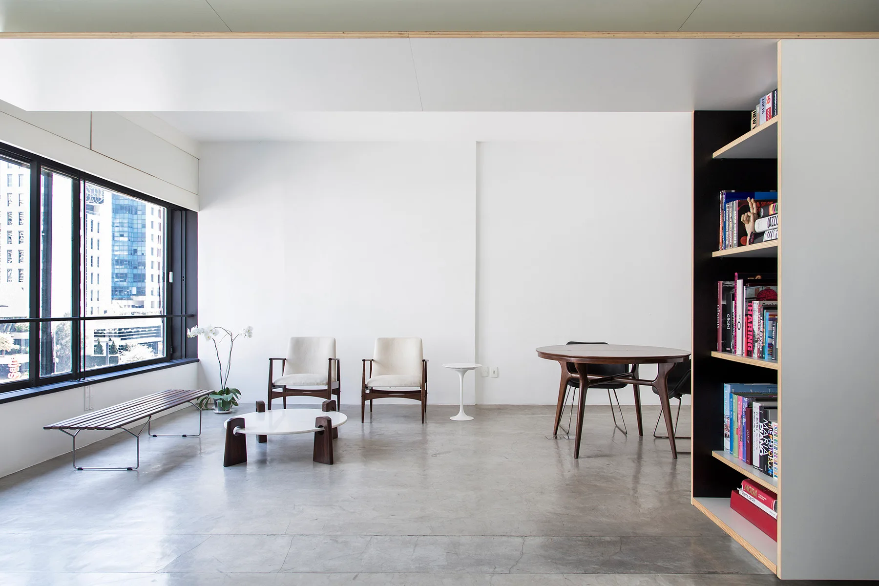 A minimalist open-plan room with concrete floors, white walls, mid-century wood chairs, a dining table, and a slatted bench under a large window.