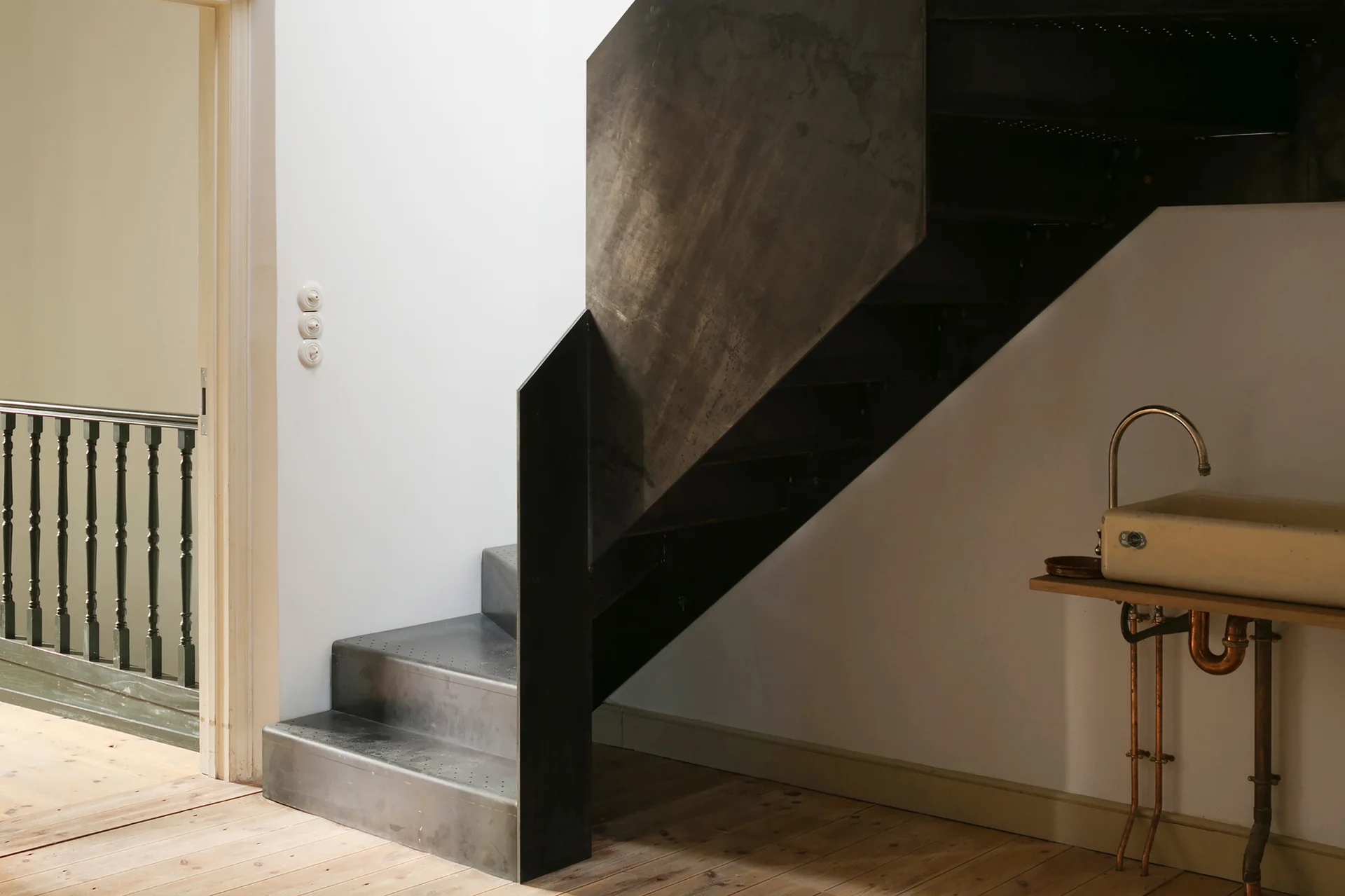 A dark industrial metal staircase with perforated steps stands next to a wall-mounted ceramic sink with exposed copper pipes on a light wood floor.