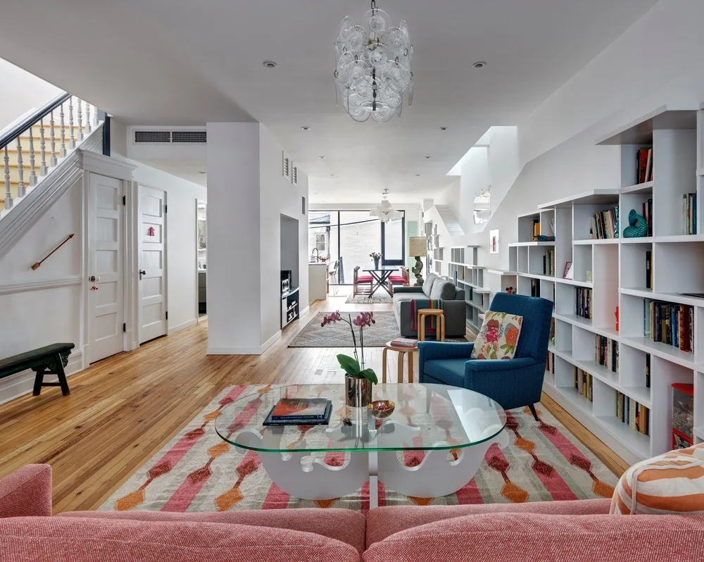 An open-concept living room features a pink sofa, a blue armchair, and extensive white bookshelves along a wall with wood floors.