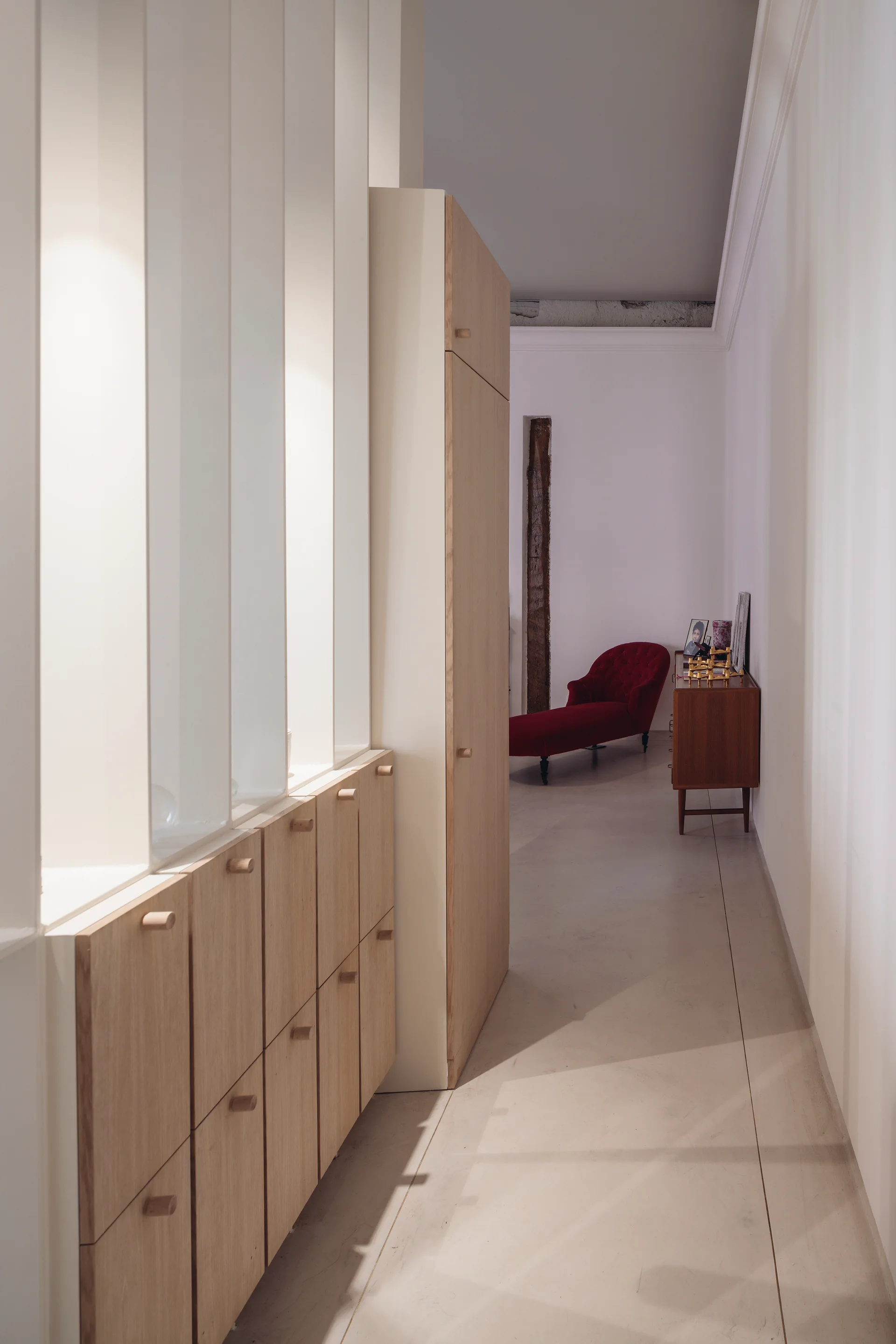 A long white hallway with low light-wood cabinets under architectural wall niches, leading to a red chaise lounge in the background.