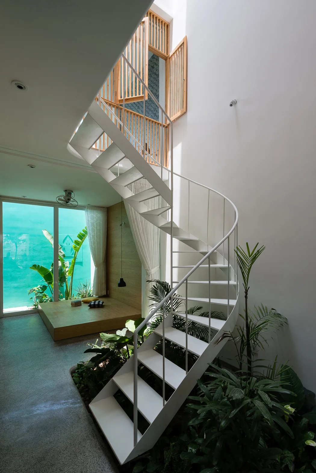A white metal spiral staircase curves over an indoor garden bed with lush green plants next to a raised wooden platform lounge area.