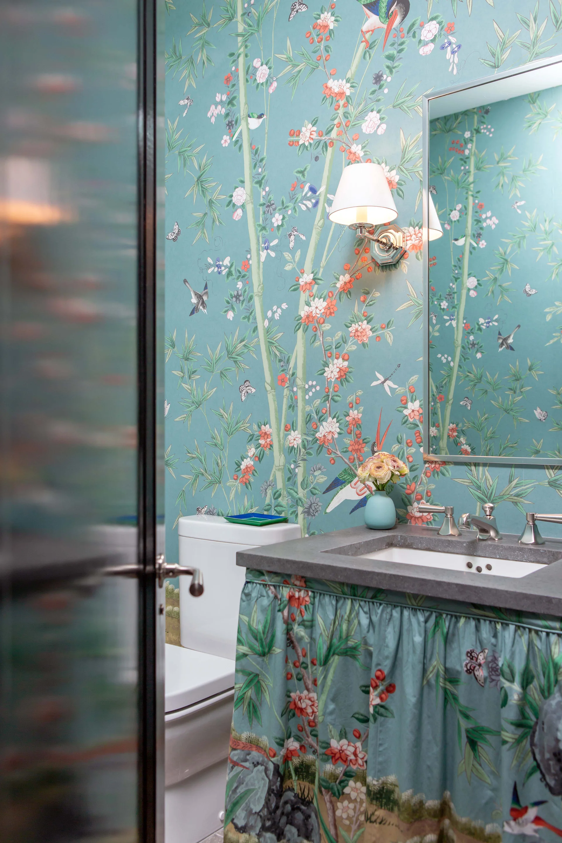 A powder room features teal Chinoiserie wallpaper, a white toilet, and a vanity with a grey stone countertop and a matching fabric skirt.