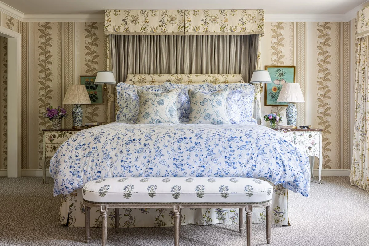 A symmetrical bedroom features a bed with blue floral linens, matching patterned wallpaper, twin bedside tables with lamps, and an upholstered bench.
