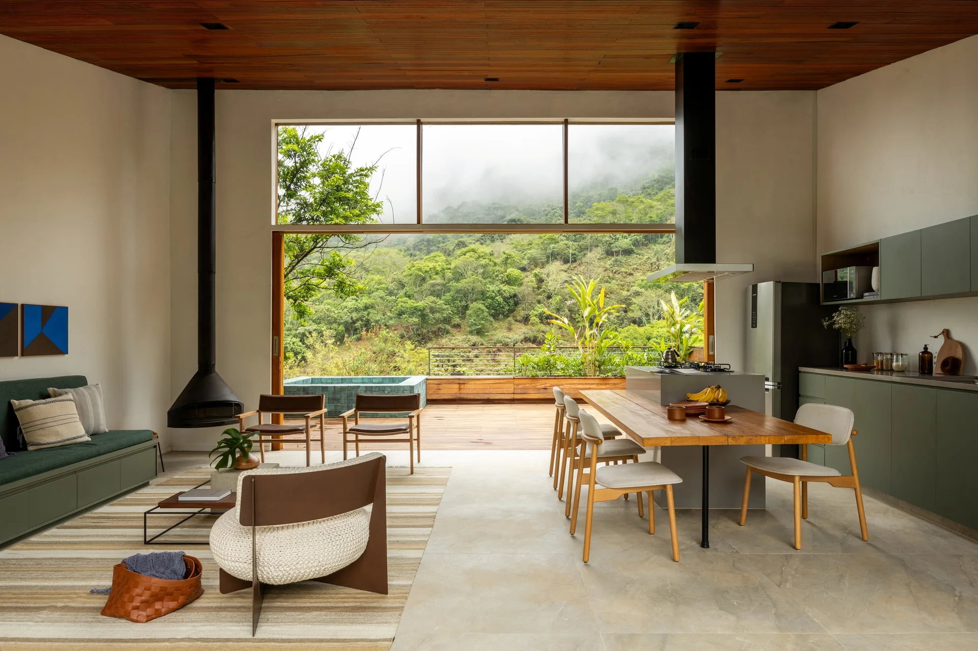 An open-concept living space features an integrated wood dining table, muted green kitchen cabinets, a suspended black fireplace, and a slatted wood ceiling.