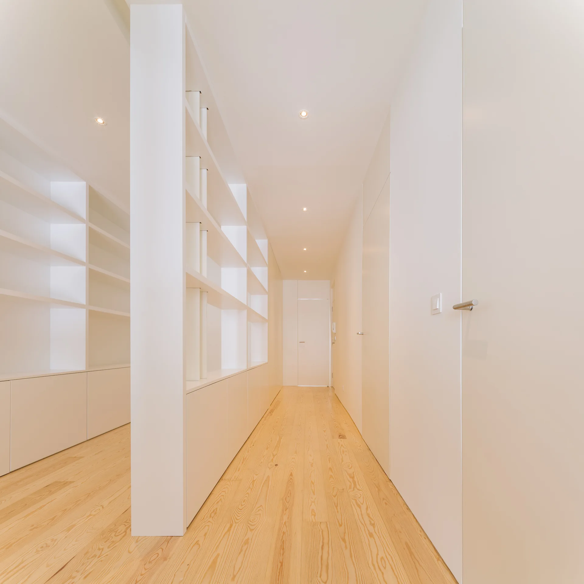 A minimalist white hallway features light wood plank flooring, flush doors, and built-in shelving with lower cabinets under recessed ceiling spotlights.