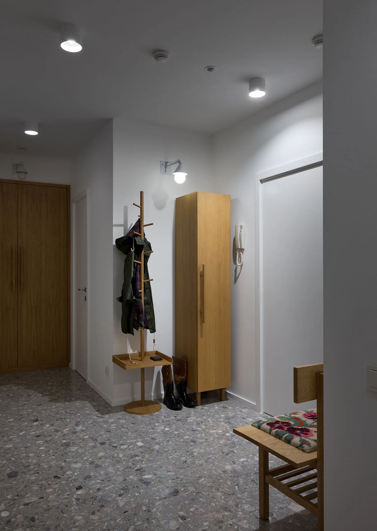 An entryway hallway features grey terrazzo flooring, a tall light wood cabinet, a matching wooden coat rack, and a bench with a floral cushion.
