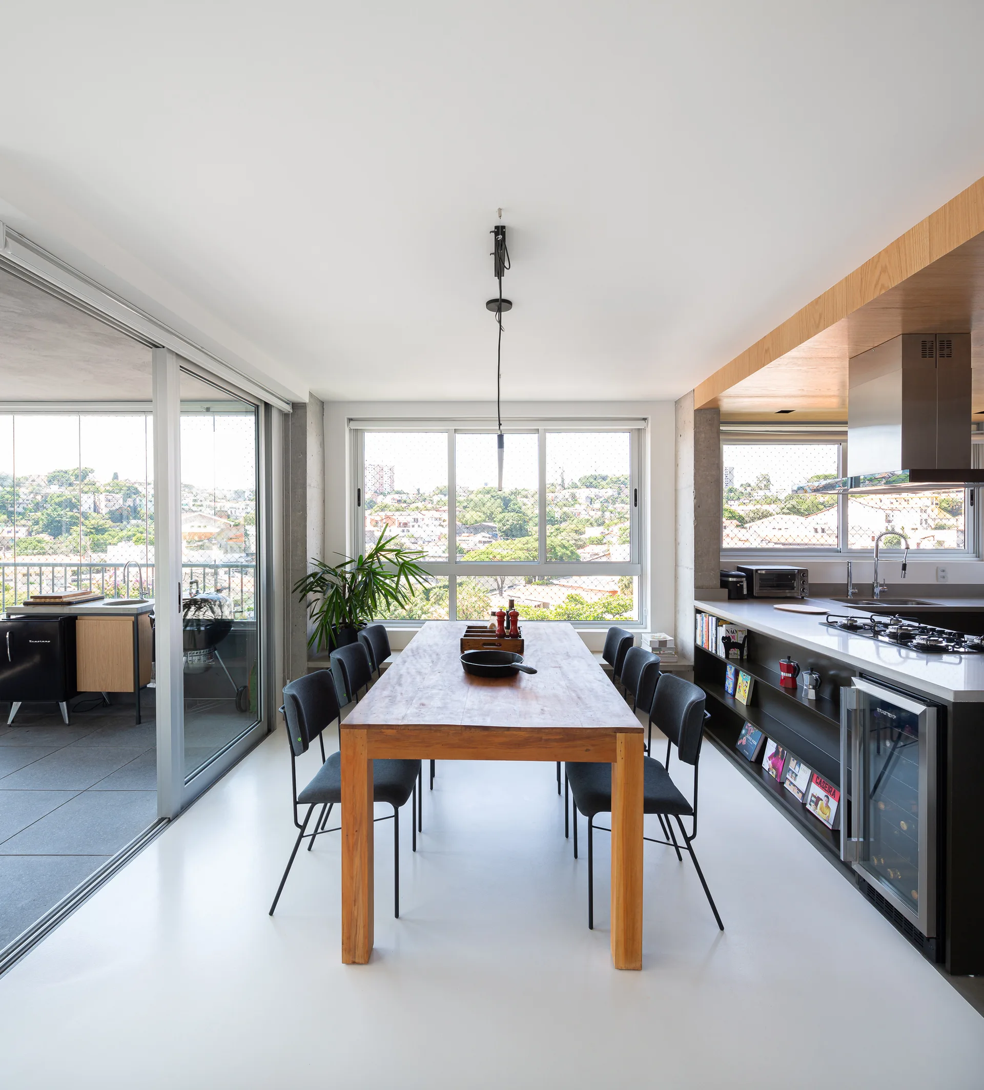 A modern open-plan dining room with a long wood table, black chairs, a seamless white floor, and glass sliding doors leading to a balcony.