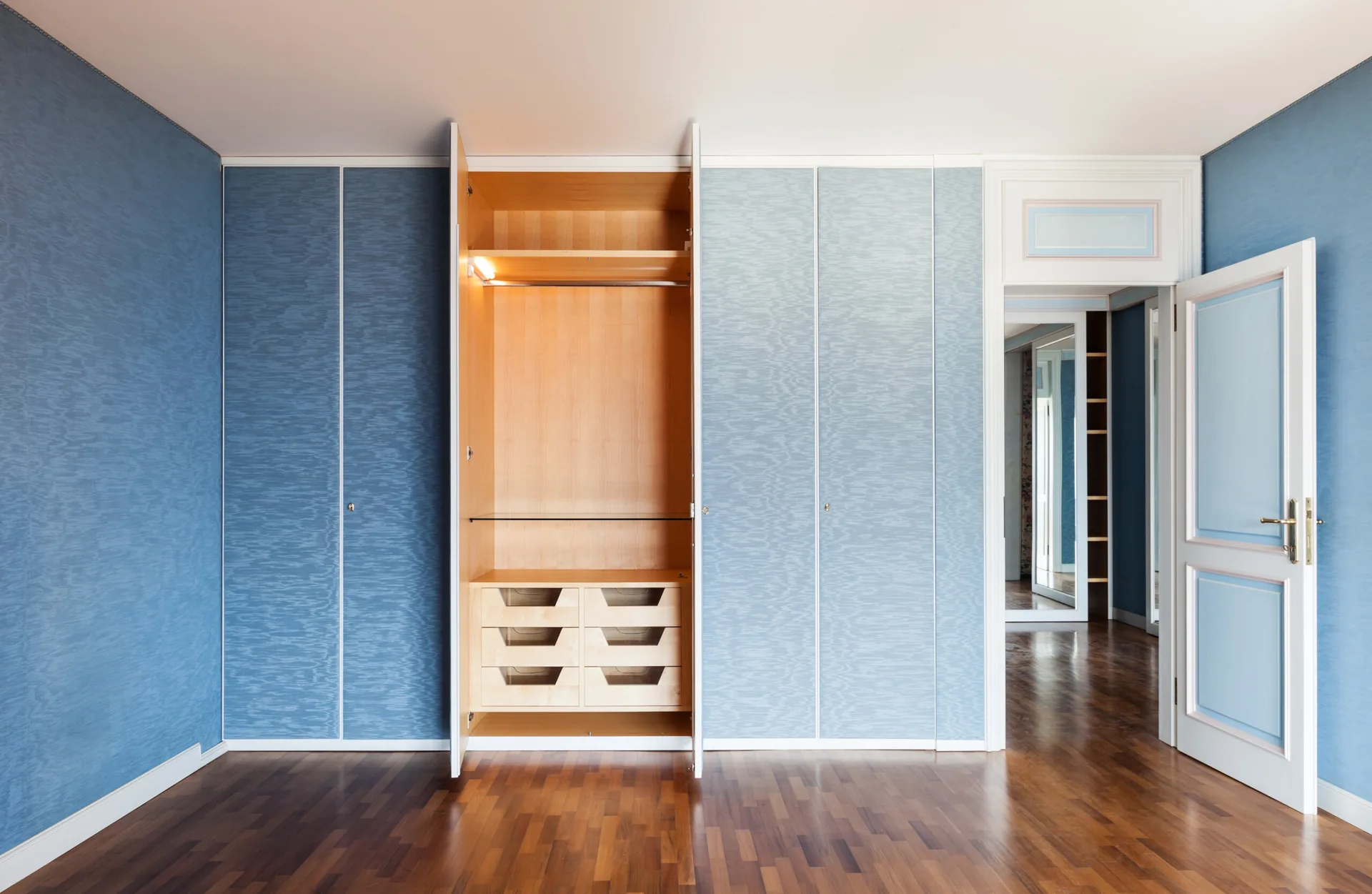 A wall of built-in closets with textured blue doors faces a dark wood floor, with one door open to show a light wood interior with drawers.