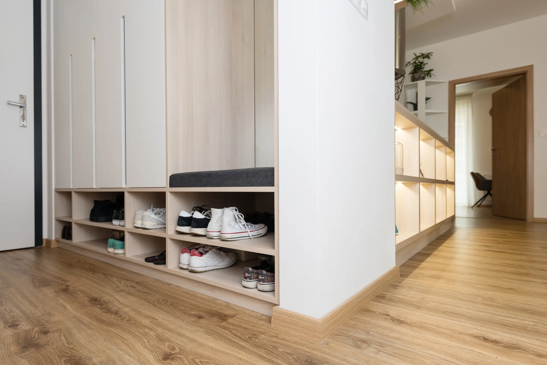 A modern entryway features custom light wood built-in cabinets with shoe storage cubbies, a grey cushioned bench, and warm accent lighting on side shelves.