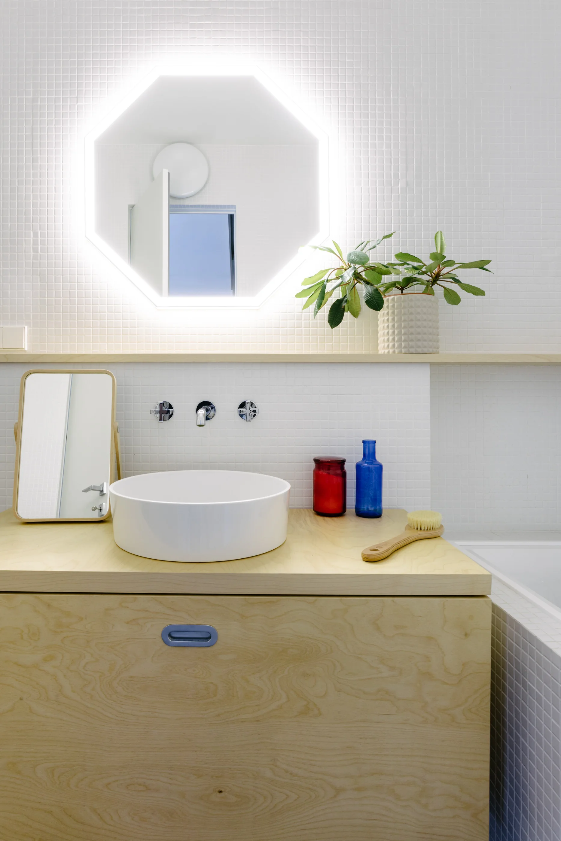 A light wood bathroom vanity with a white vessel sink stands against a white square-tiled wall beneath a glowing backlit octagonal mirror.