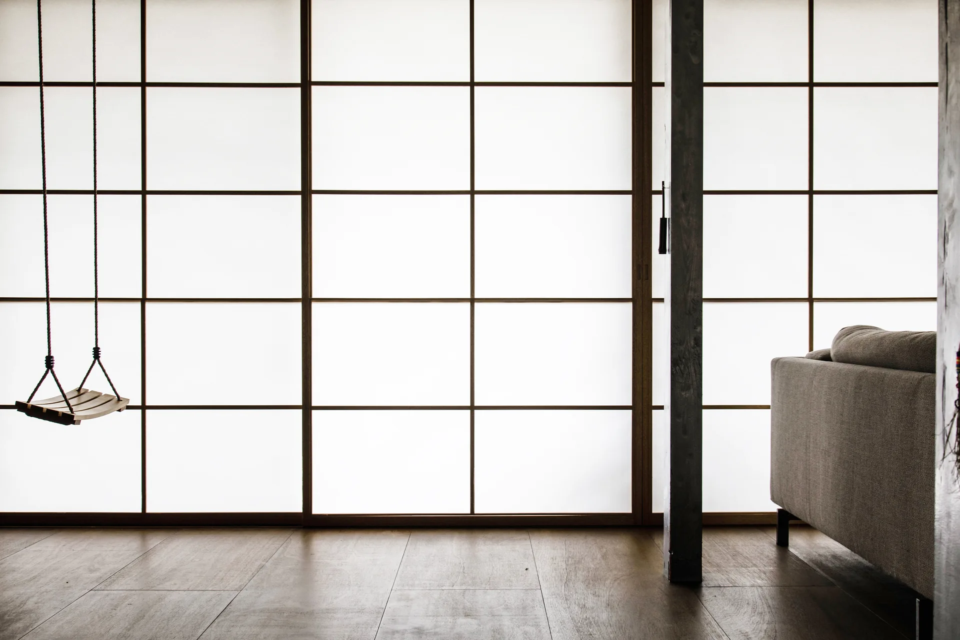 A minimalist room featuring a backlit shoji screen wall, a wooden hanging swing, a dark support post, and a grey fabric sofa on wood flooring.
