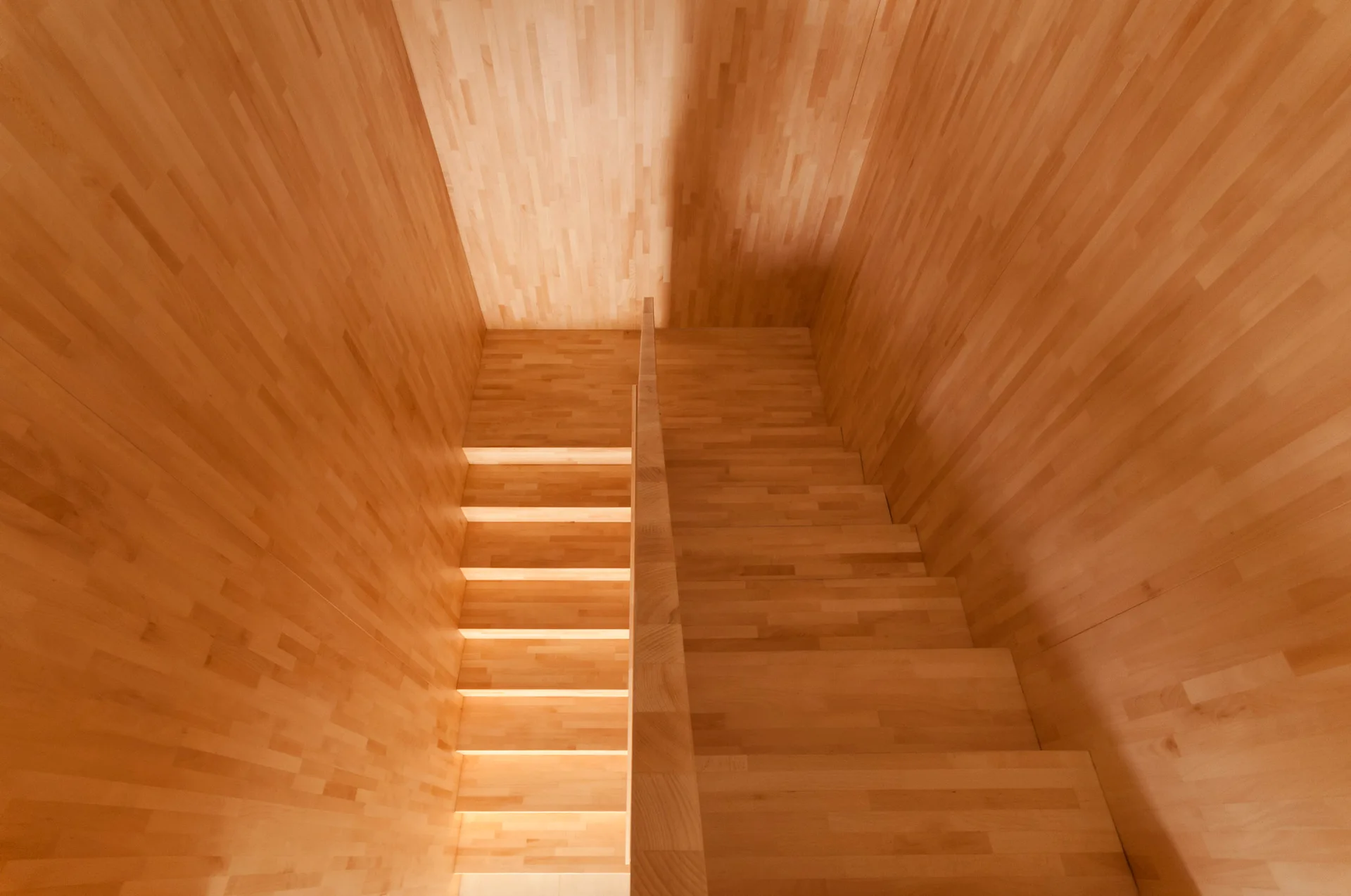 A top-down view of a modern staircase constructed from light wood with matching wood paneling on the surrounding walls and a central wooden handrail divider.