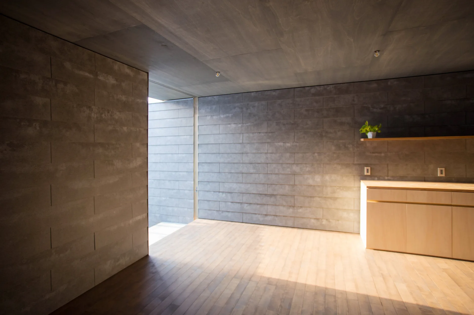 A minimalist room featuring grey textured tile walls, light wood flooring, a simple wooden cabinet, and natural sunlight coming from an open doorway.