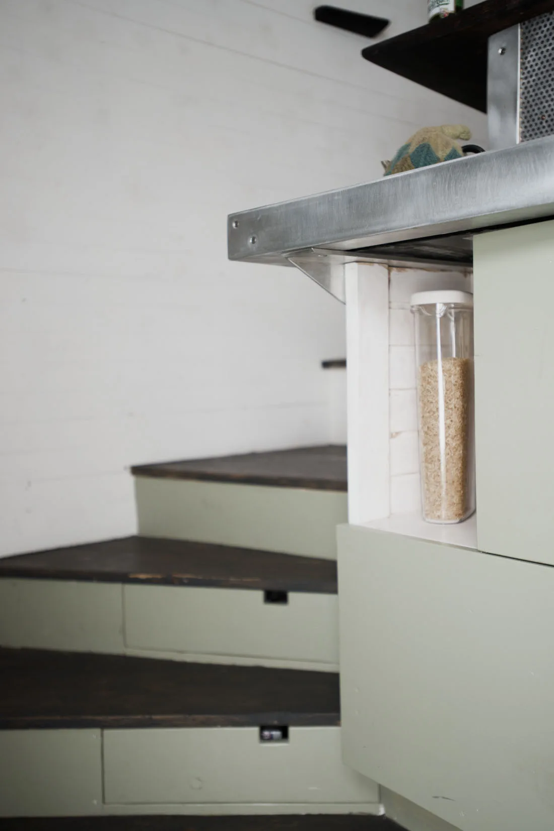 A curved wooden staircase with sage green risers and integrated drawers stands next to a kitchen cabinet with a metal countertop.