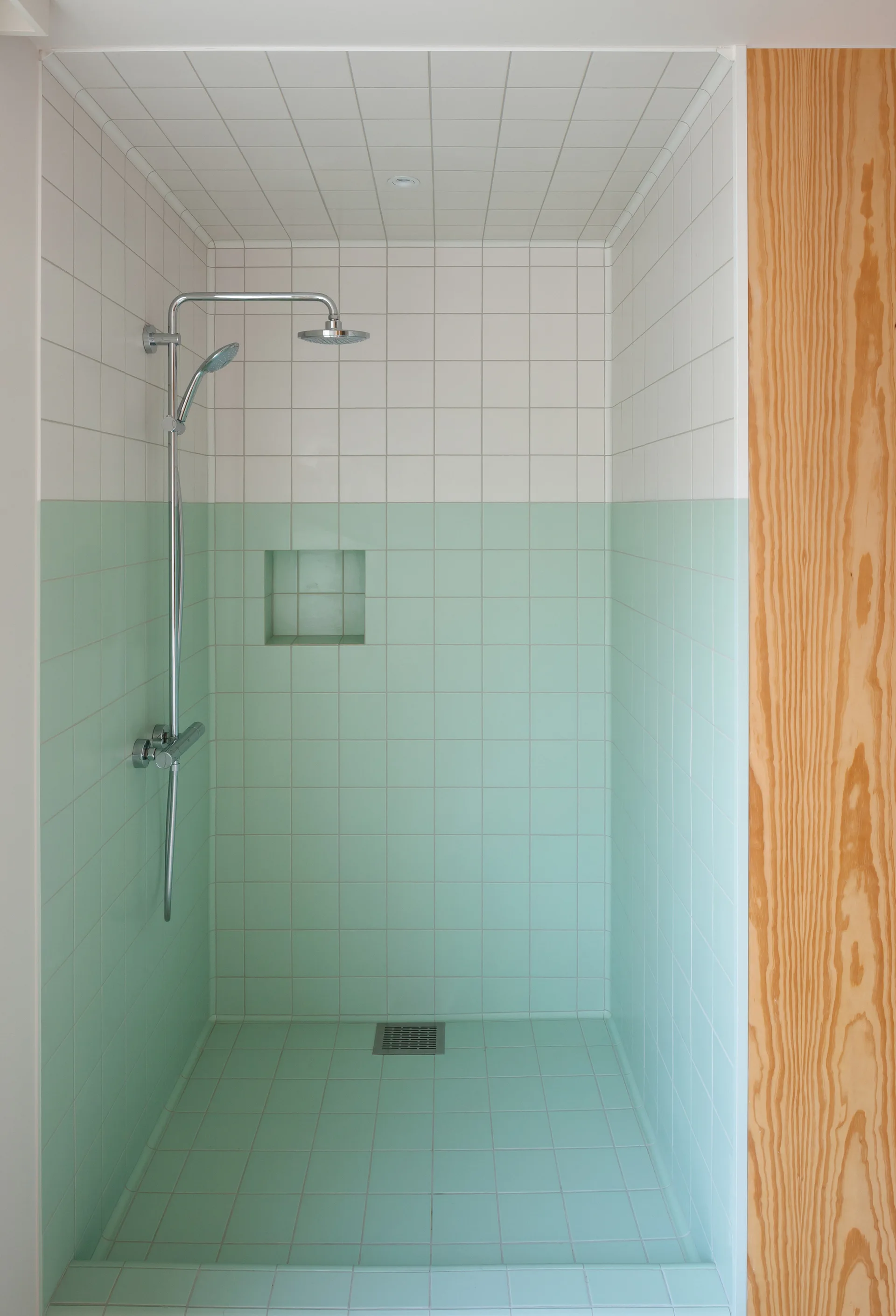 A minimalist shower stall features a two-tone grid of mint green and white square tiles, a recessed wall niche, and a vertical wood panel.