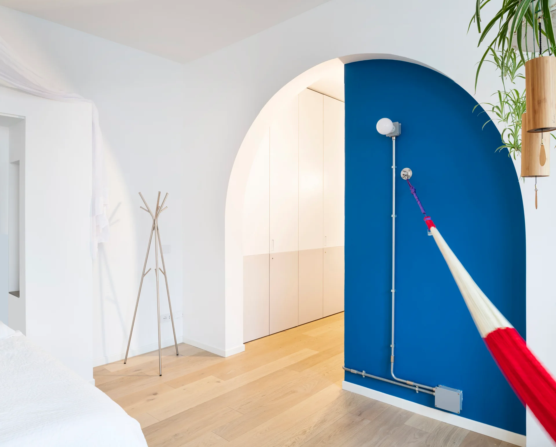 A minimalist white bedroom features a cobalt blue arched wall section with exposed metal conduit, light wood floors, and a metal coat rack.