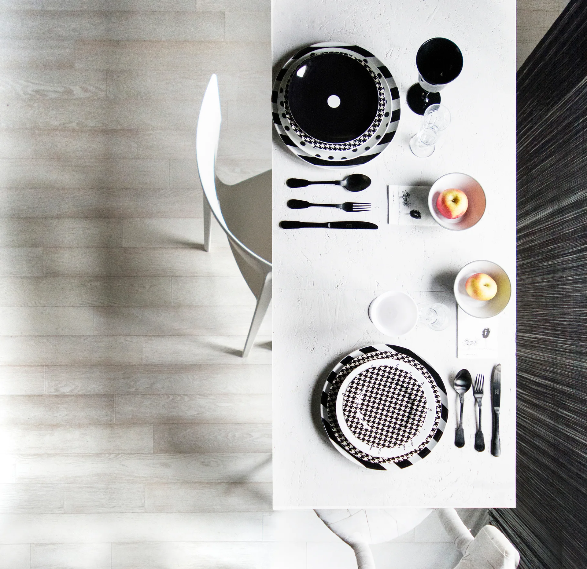 A top-down view of a white dining table set with graphic black and white patterned plates, black cutlery, and bowls containing fresh apples.