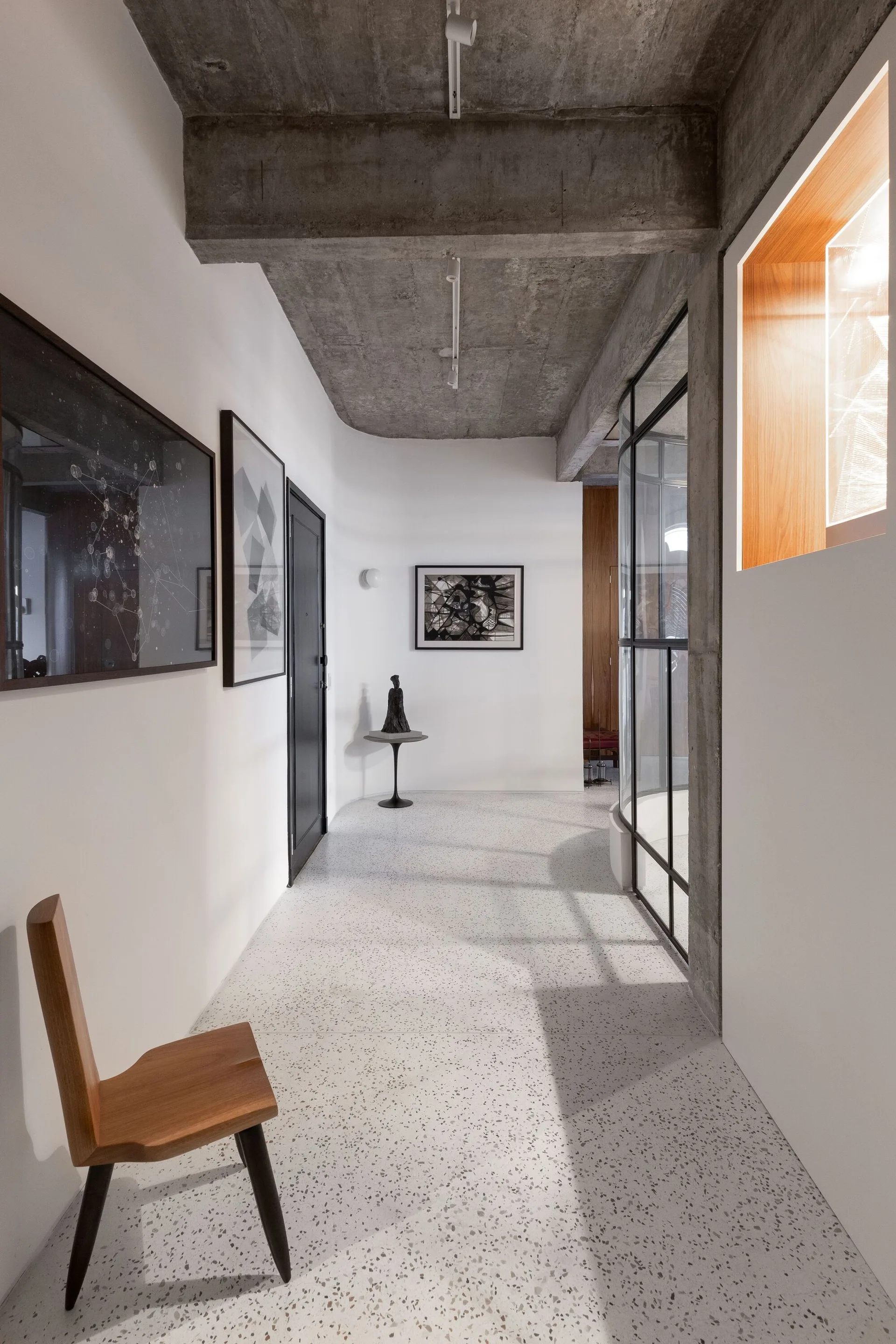 A minimalist residential hallway features a raw concrete ceiling, terrazzo flooring, white walls with framed art, and a wooden chair.