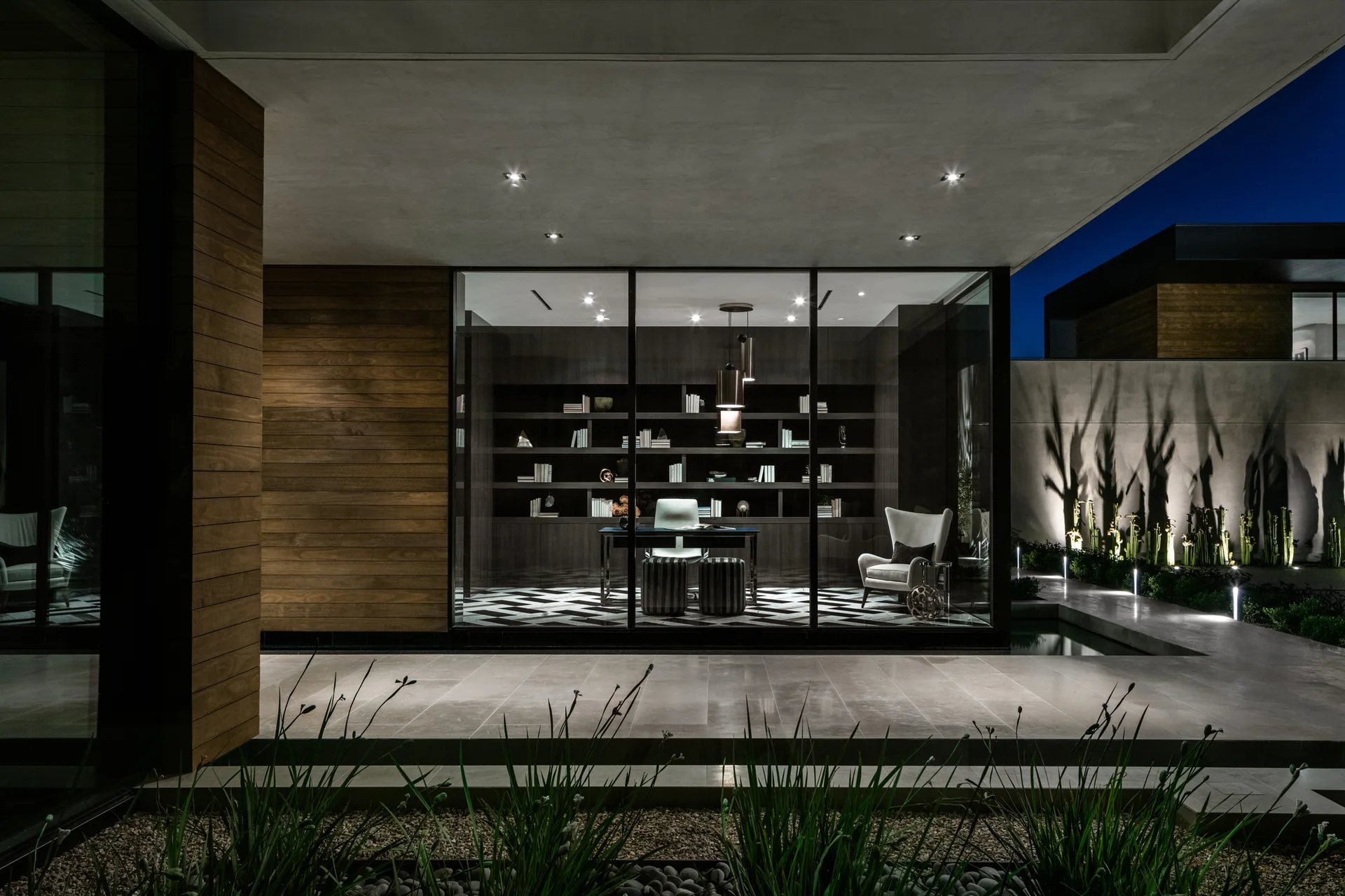 A modern home office with a dark built-in bookcase and chevron flooring is visible through a large glass wall from a lighted patio at night.