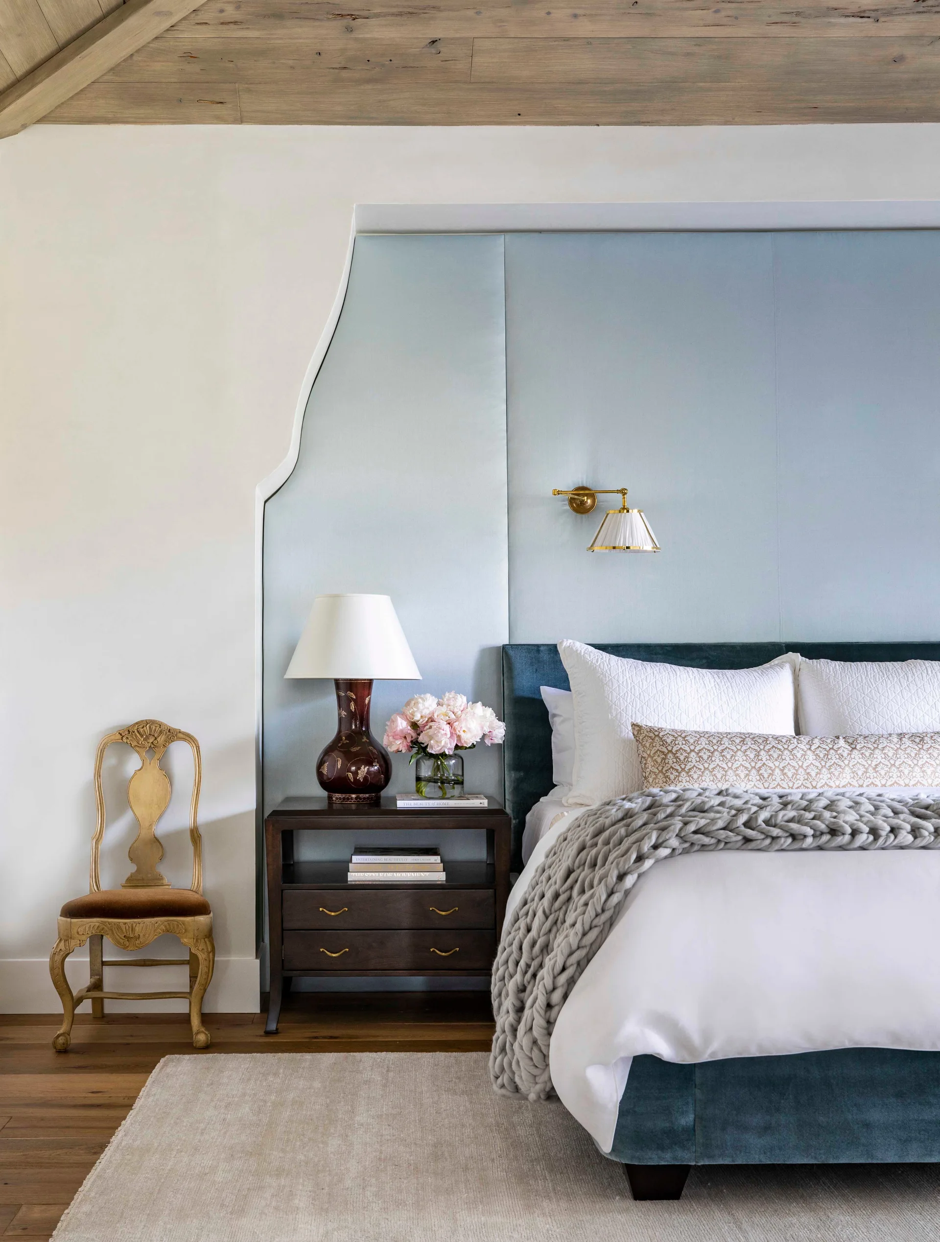 A bedroom featuring an upholstered blue headboard, a light blue fabric wall panel, a dark wood nightstand, and an ornate gilded chair.