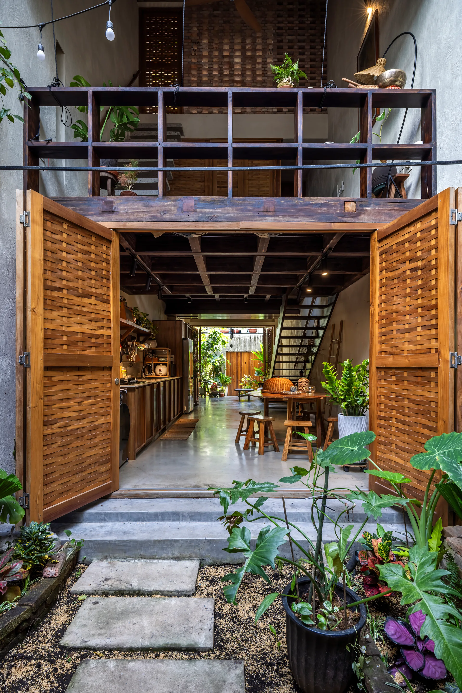 A view from a garden looking into a tropical open-concept kitchen with concrete floors, woven wooden doors, and an overhead wooden loft structure.