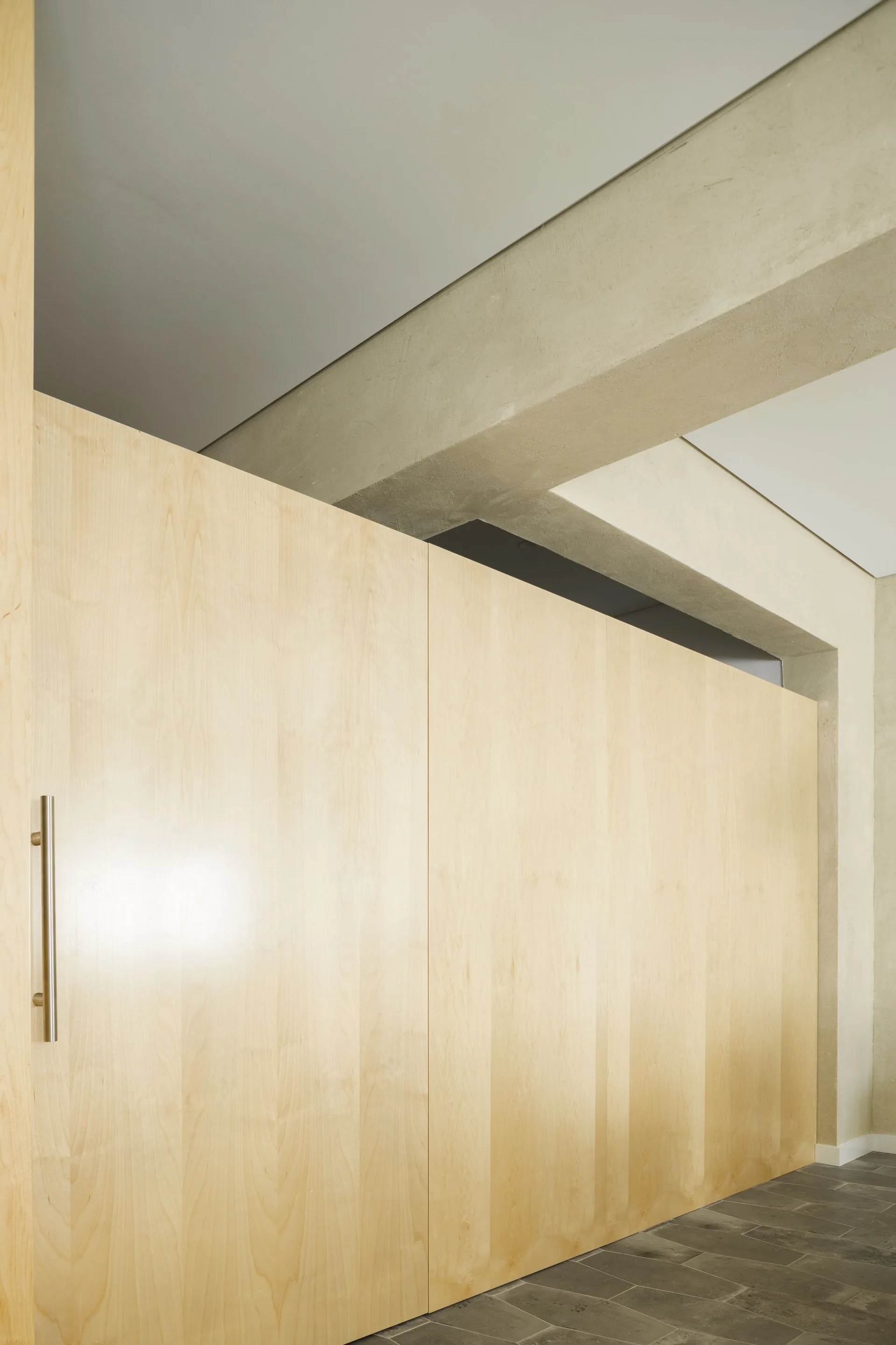 Light-toned wood cabinetry with a silver vertical handle stands beneath an exposed concrete ceiling beam and gray tiled flooring.