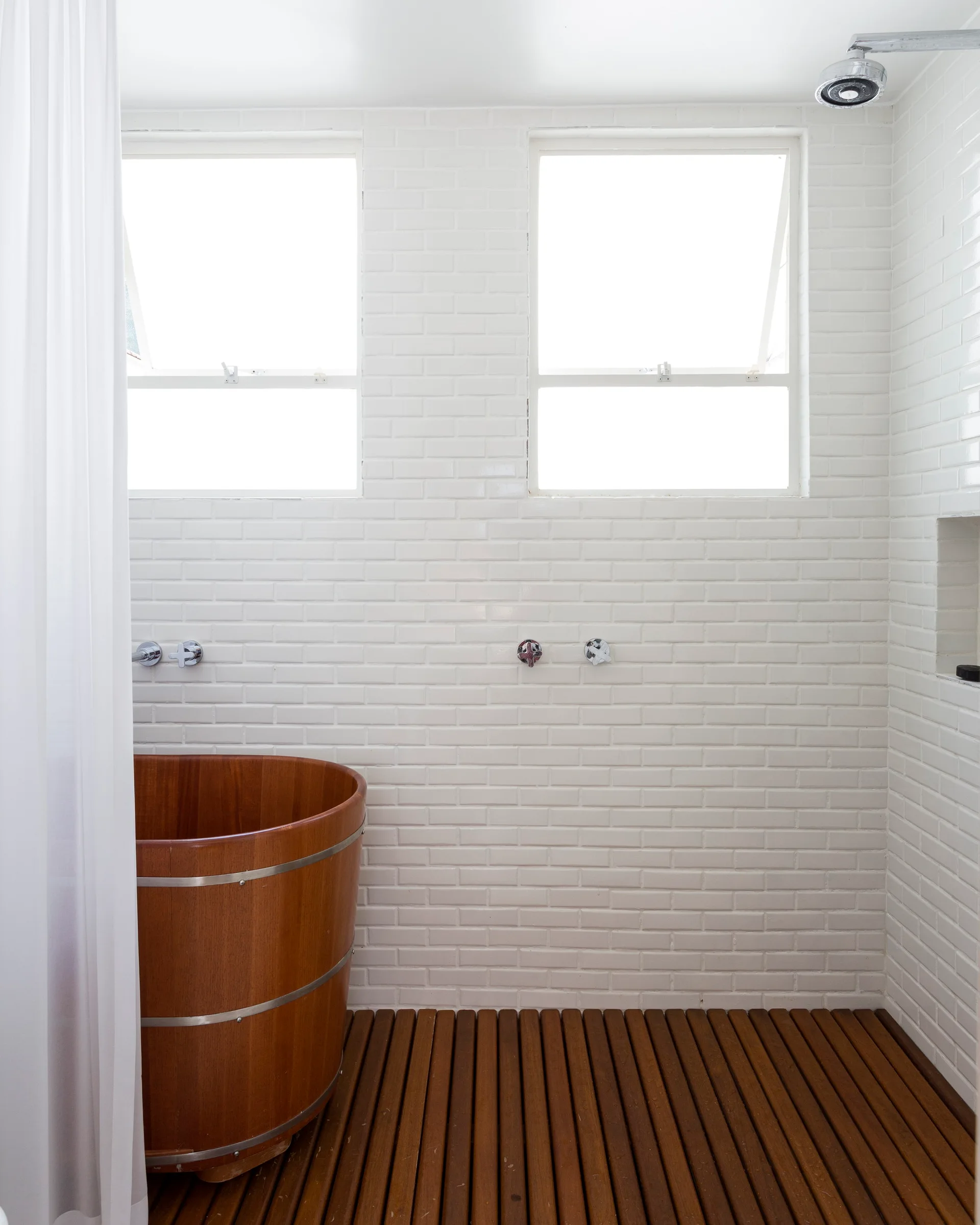 A bathroom shower area featuring a cylindrical wooden soaking tub on a slatted wood floor against white subway tiled walls with two windows.