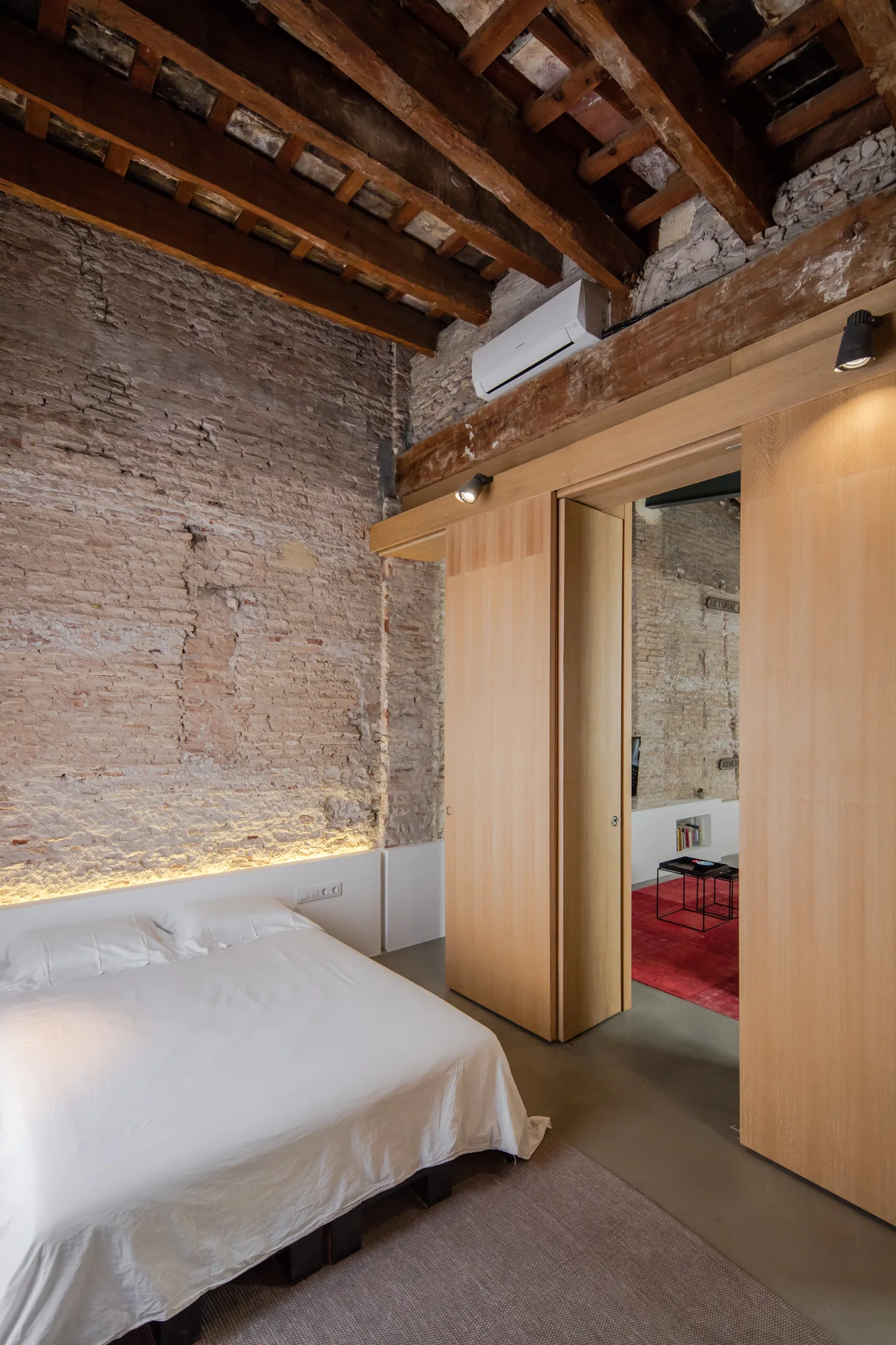 A minimalist bedroom featuring a bed with white linens against an exposed brick wall under a rustic wood-beamed ceiling with a sliding wood door.