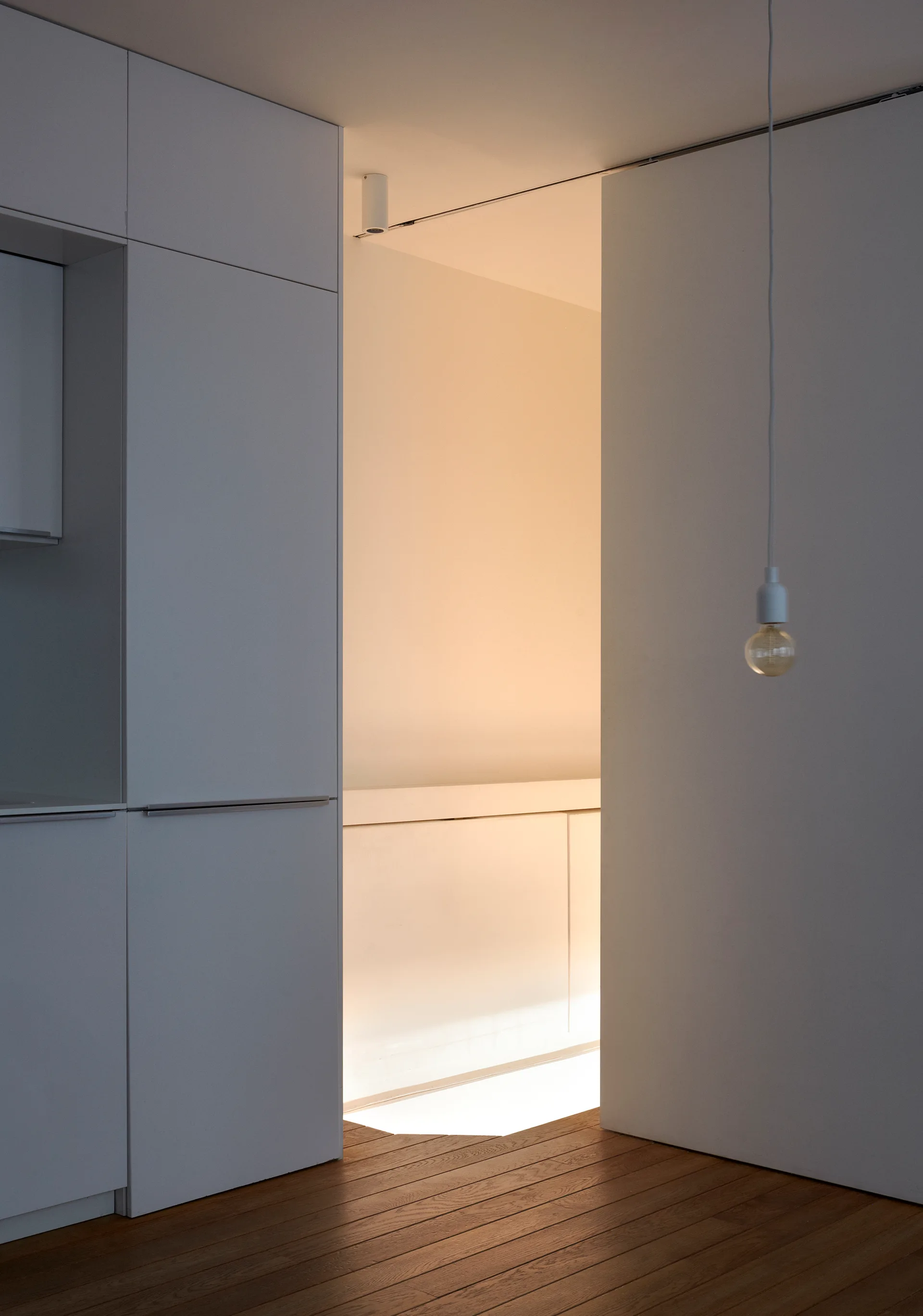 A minimalist hallway featuring white built-in handleless cabinets, wood plank flooring, a doorway with warm interior light, and a single hanging bulb.