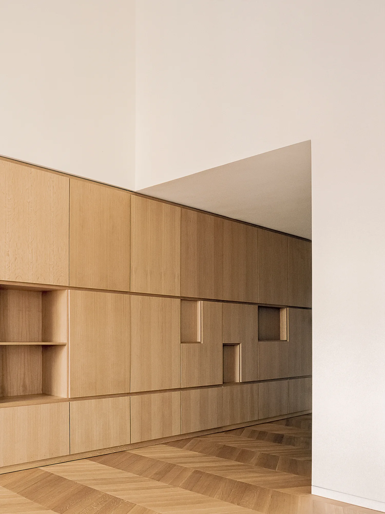 A minimalist hallway features a wall of light wood built-in cabinets with recessed niches above a herringbone pattern wood floor.