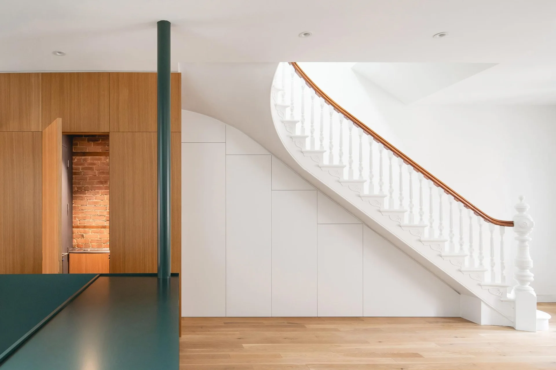 A white curved staircase features built-in under-stair storage panels alongside a wood-paneled wall, exposed brick accent, and light wood floors.