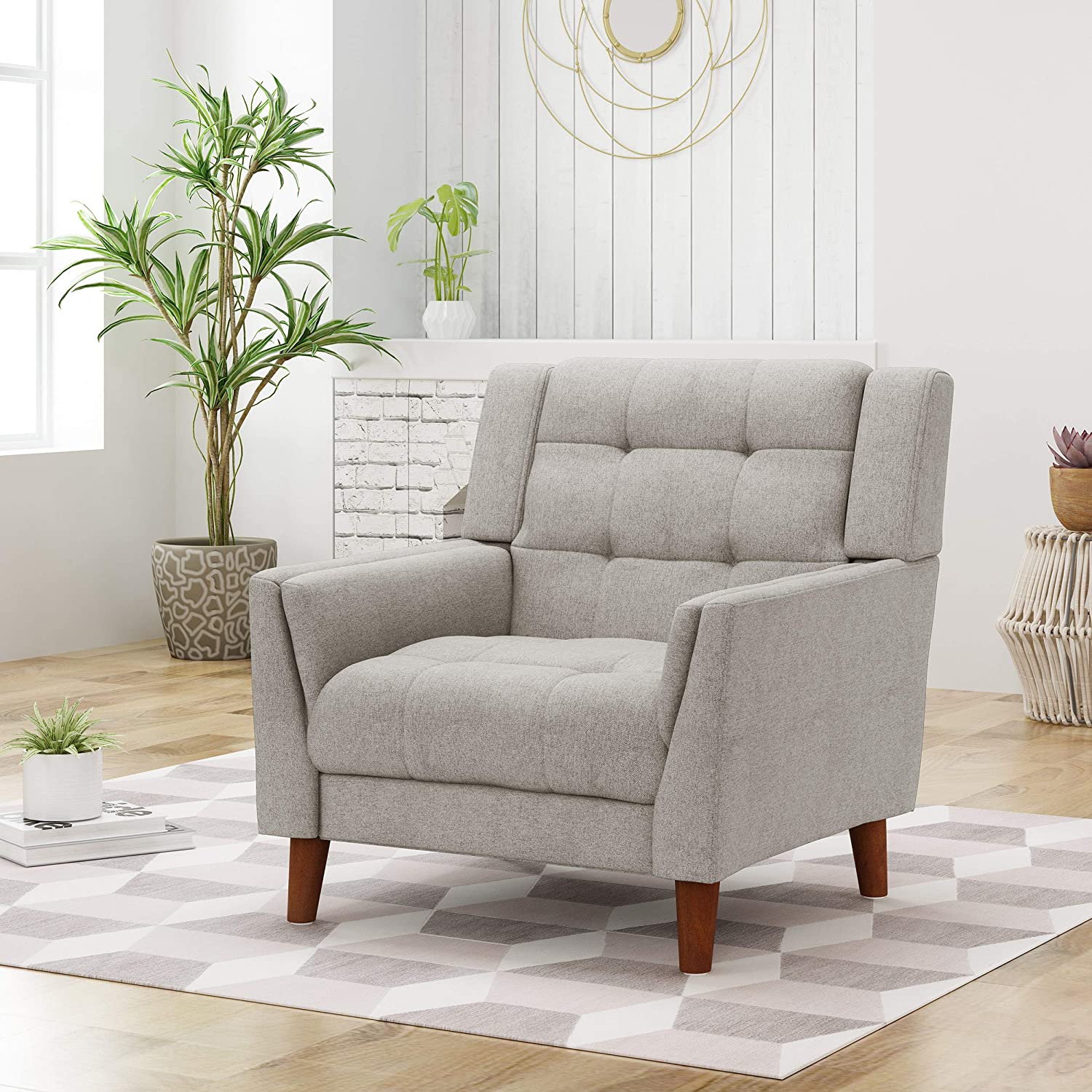A light gray tufted armchair with tapered wooden legs sits on a geometric patterned area rug next to potted plants.