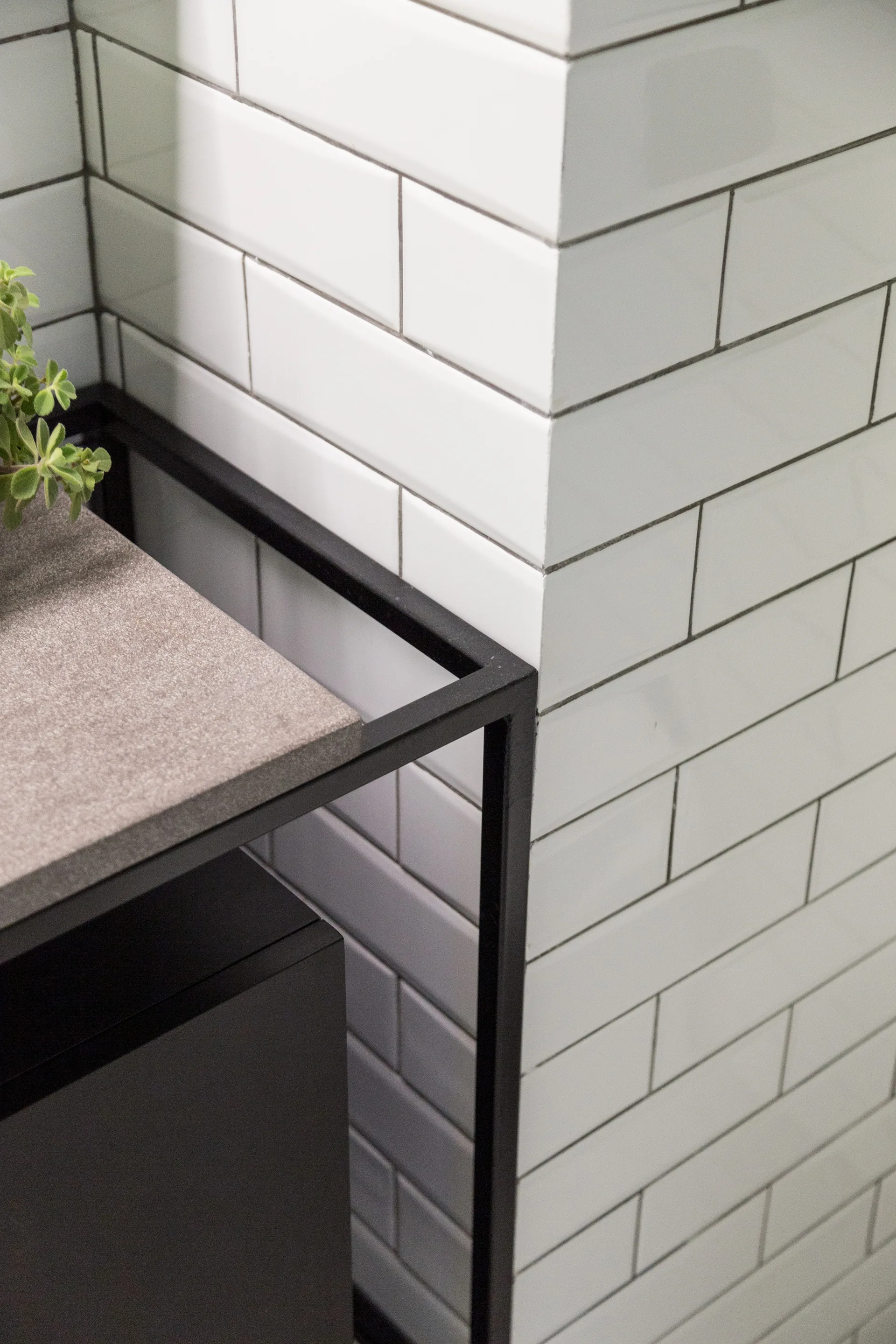 A close-up of a bathroom corner showing a black metal frame vanity with a stone countertop against a white subway tile wall with dark grout lines.