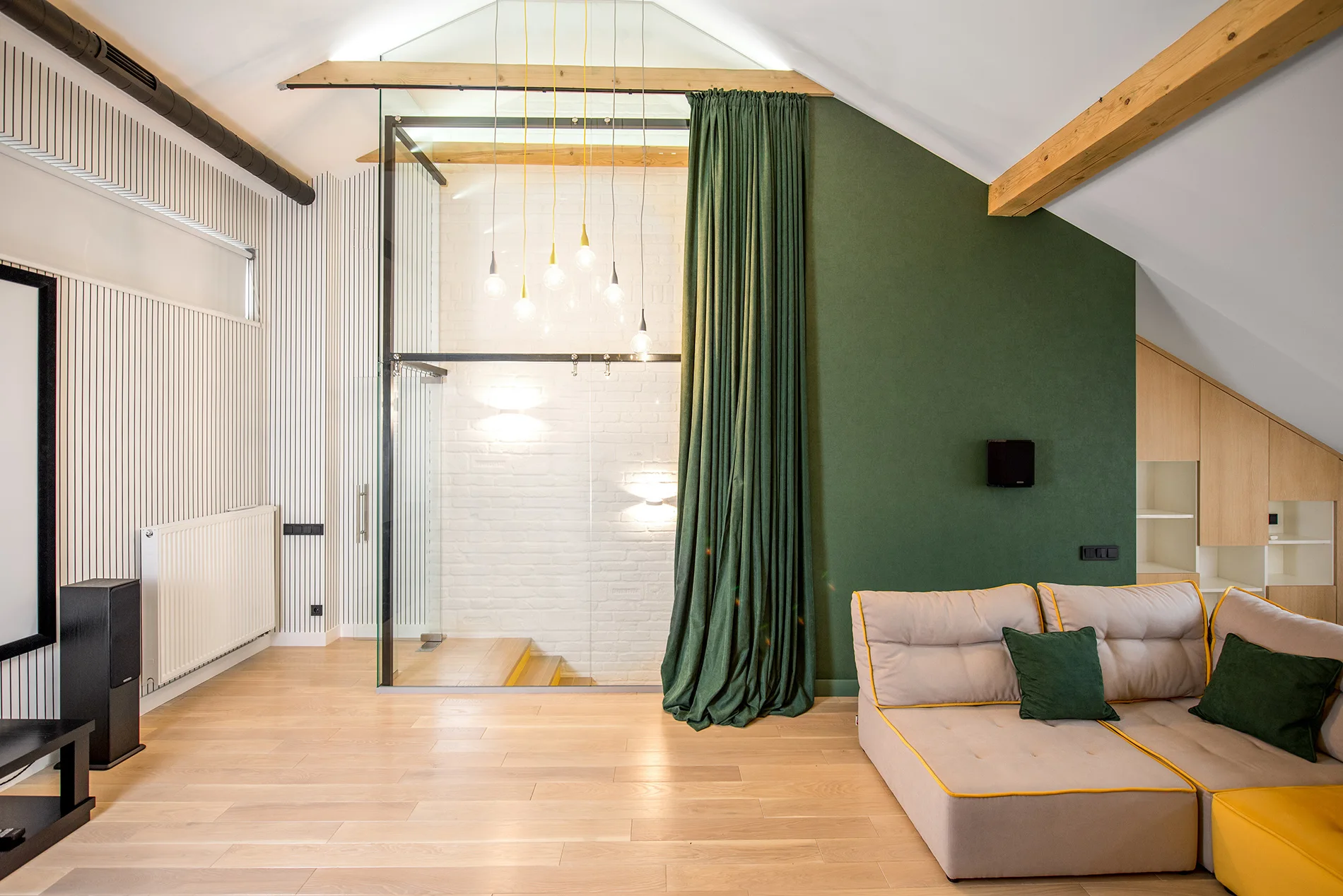 A modern loft living area features a modular beige sofa, a bold forest green accent wall, and a glass room partition under a vaulted wood-beam ceiling.