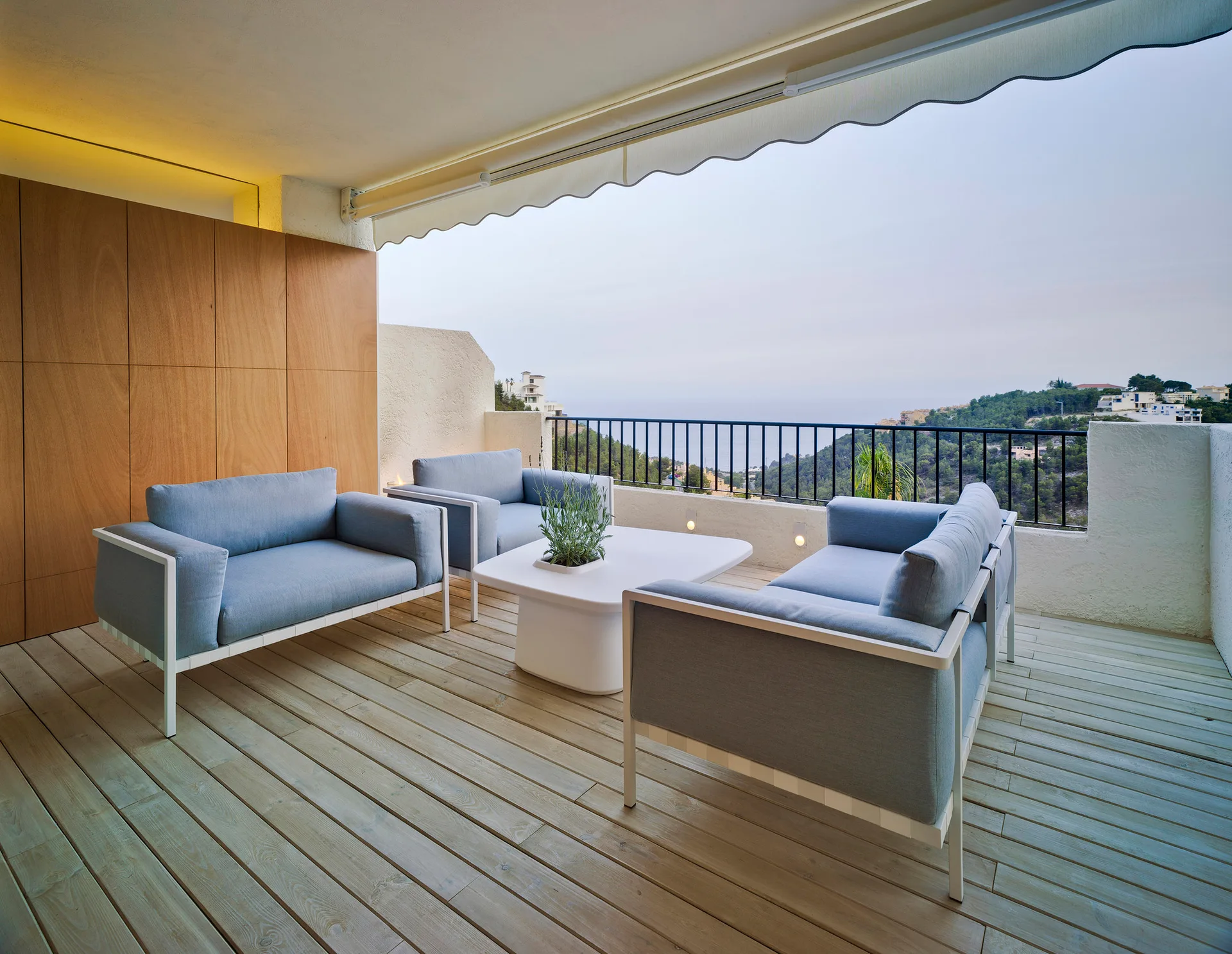 Blue outdoor sofas and a white coffee table sit on a wood balcony deck with a black railing overlooking a coastal landscape.