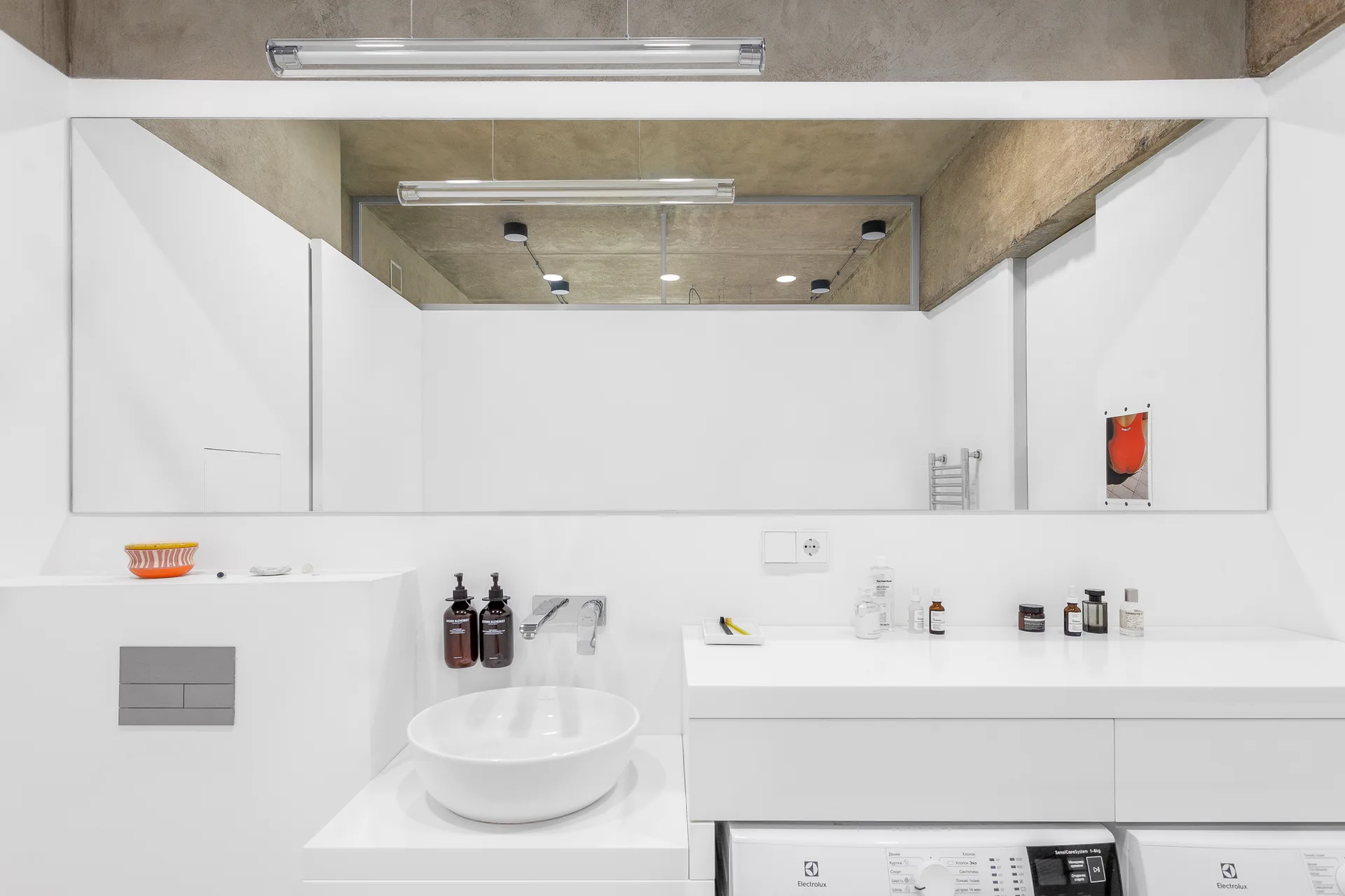 A minimalist white bathroom features a round vessel sink, a large rectangular mirror, and a washing machine integrated beneath the vanity.