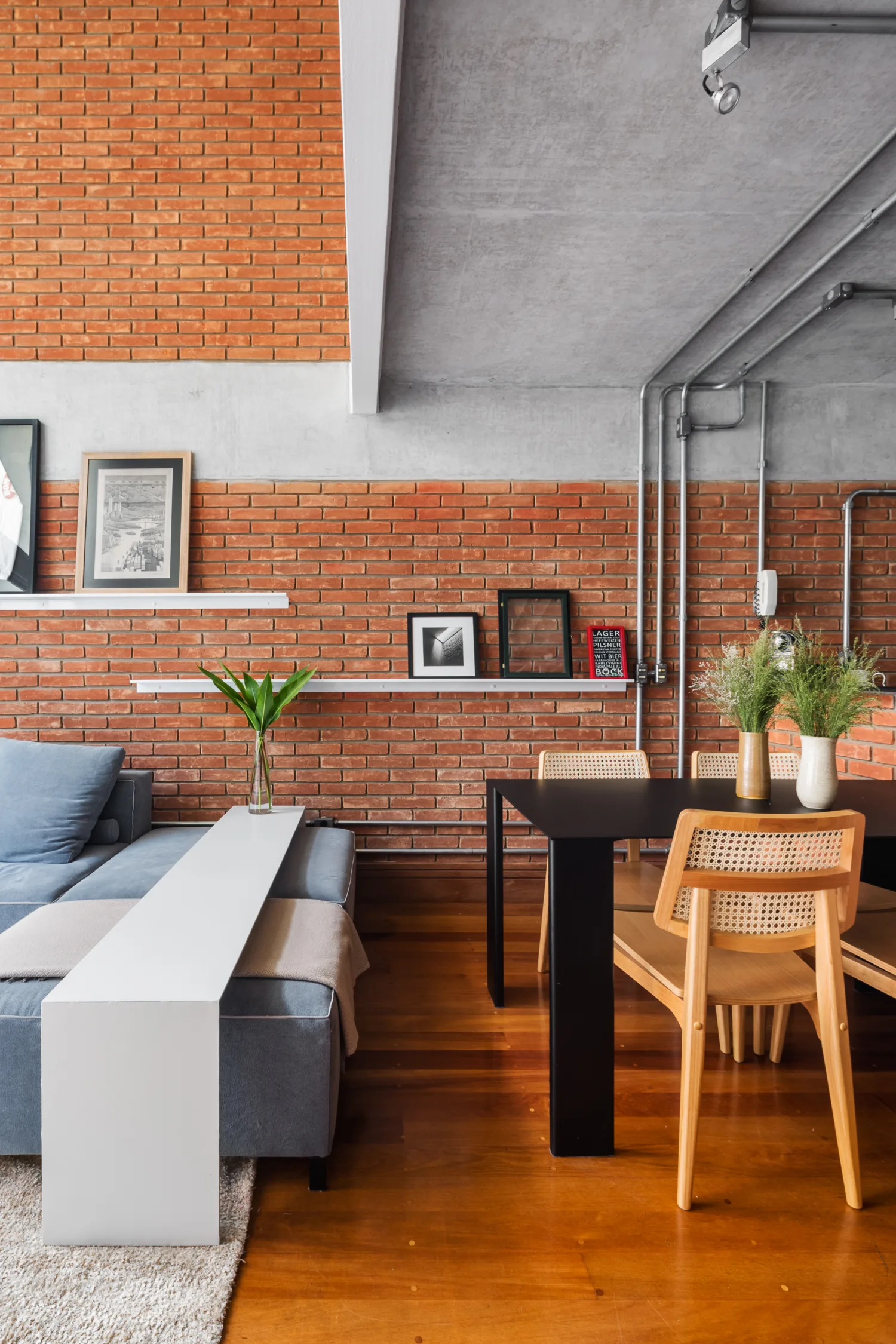 An industrial open-plan living and dining area featuring an exposed brick wall, a black dining table with cane chairs, and a grey sectional sofa.