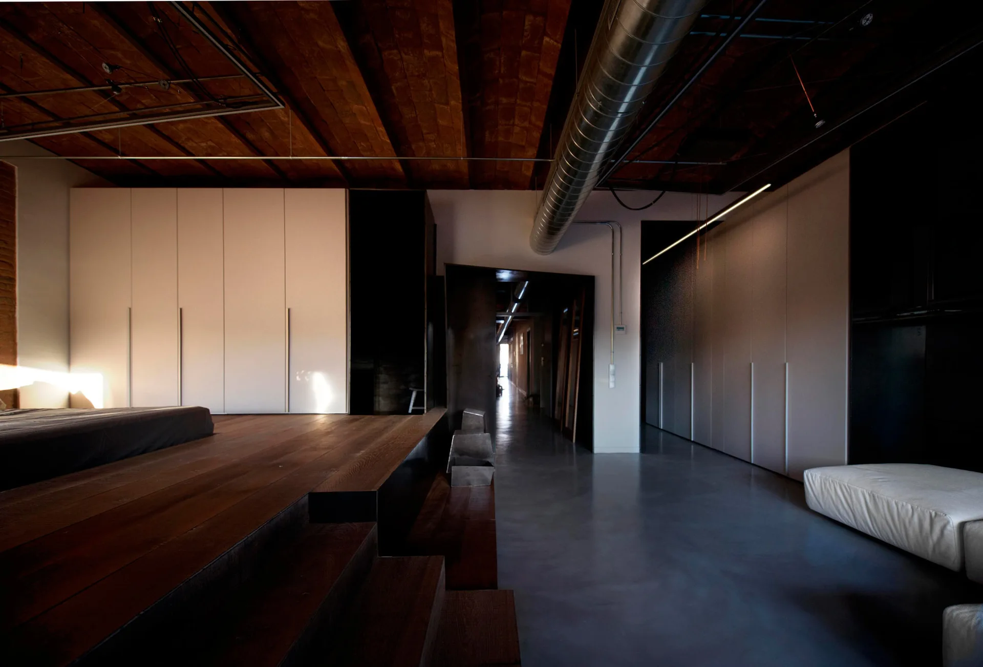 An industrial loft bedroom features a raised dark wood platform, seamless light gray wardrobes, and an exposed brick ceiling.