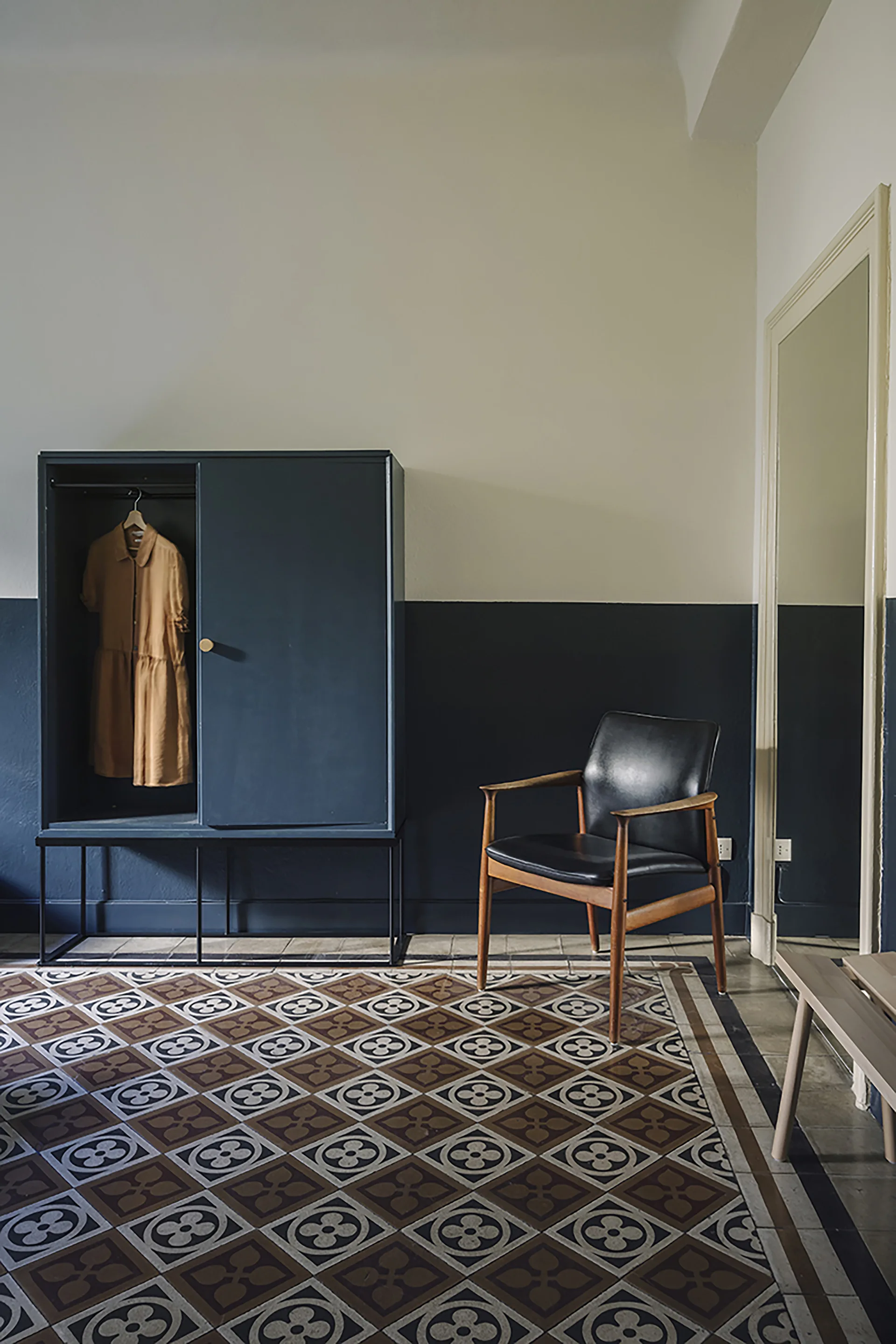 A dark blue wardrobe and a black leather armchair sit against a half-painted navy and white wall on patterned geometric floor tiles.