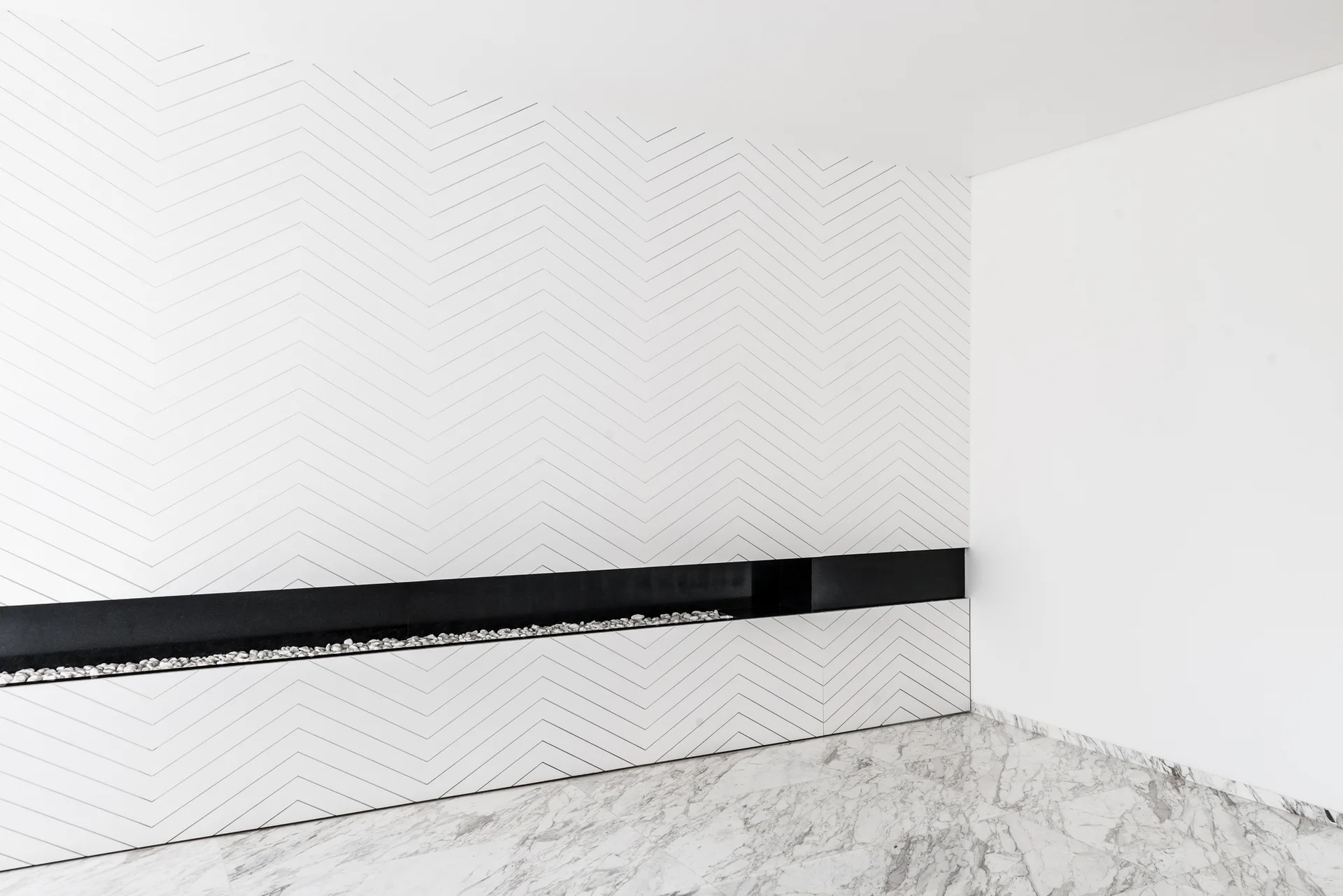A minimalist fireplace features a long horizontal black inset with white stones, set into a white chevron-patterned wall above polished marble flooring.