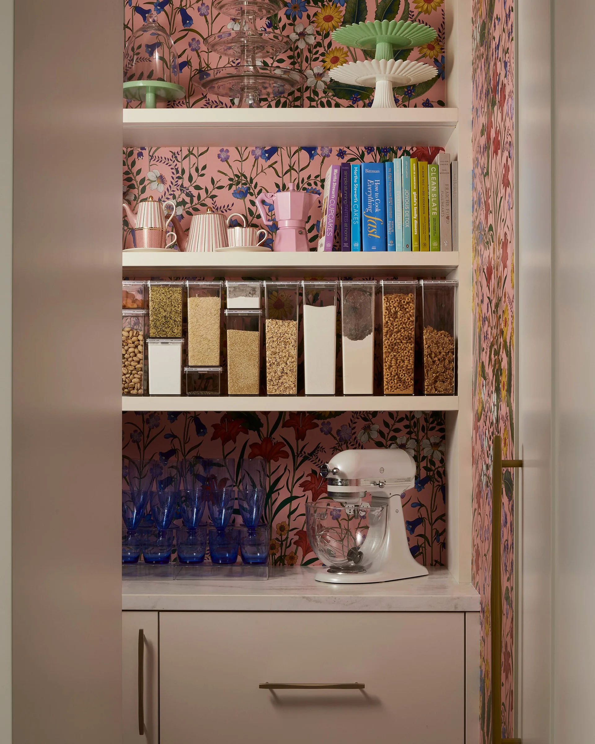 White pantry shelves filled with clear dry-food containers, books, and kitchen accessories against a pink floral wallpaper backdrop.